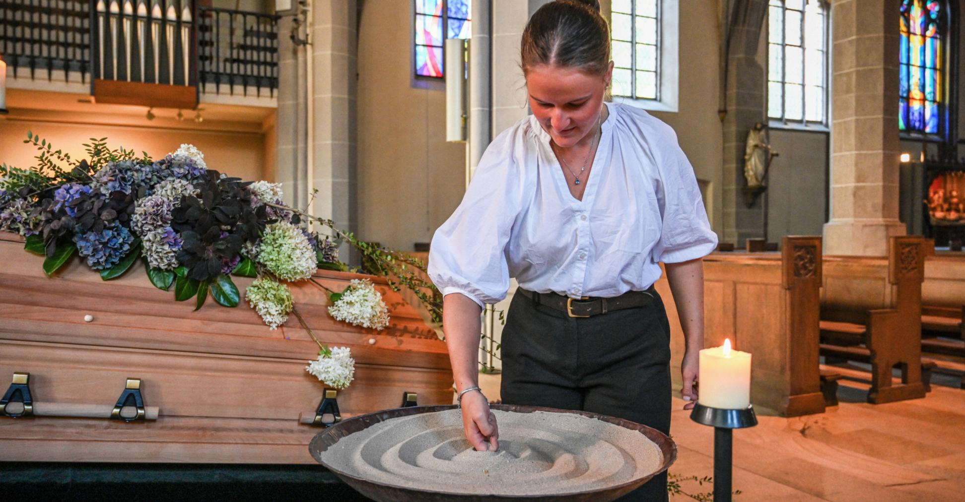 Eine Bestattering formt Sand in einer Schale, die vor einem geschmückten Sarg steht.