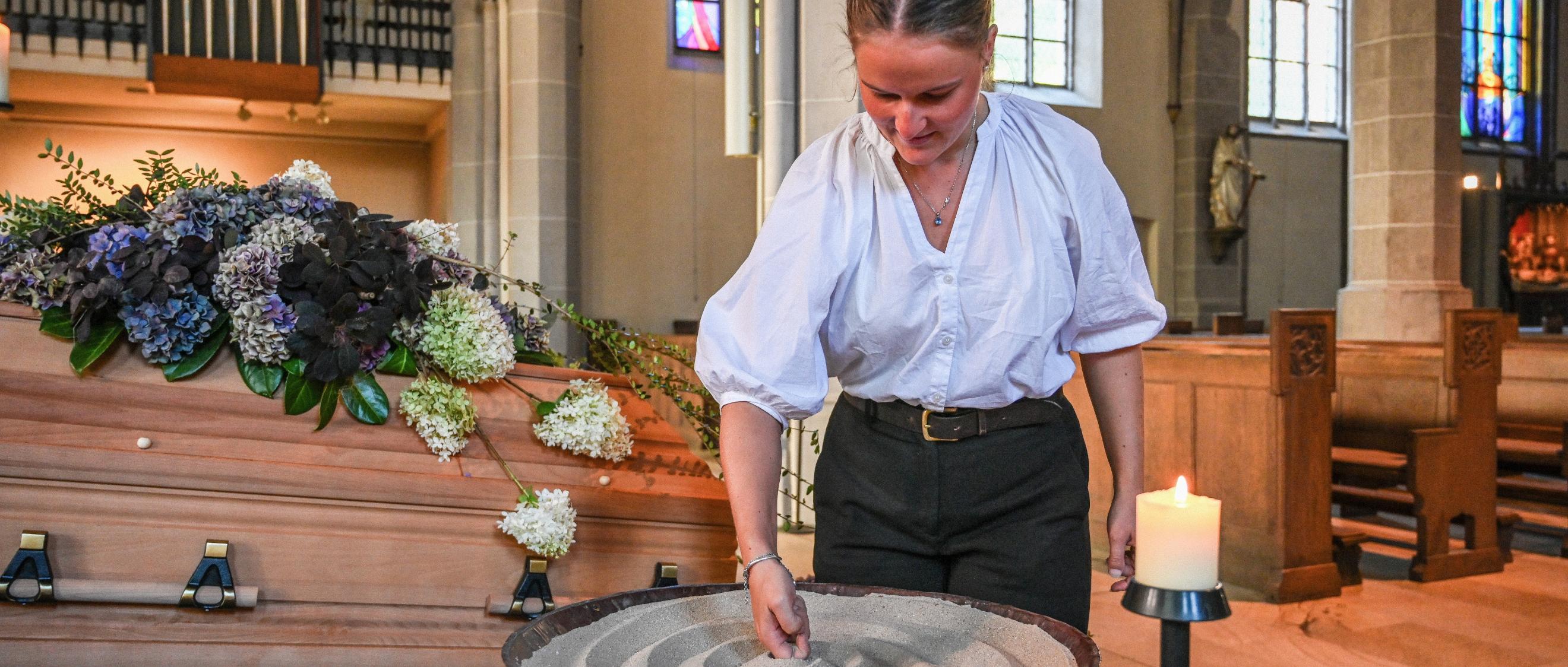 Eine Bestattering formt Sand in einer Schale, die vor einem geschmückten Sarg steht.