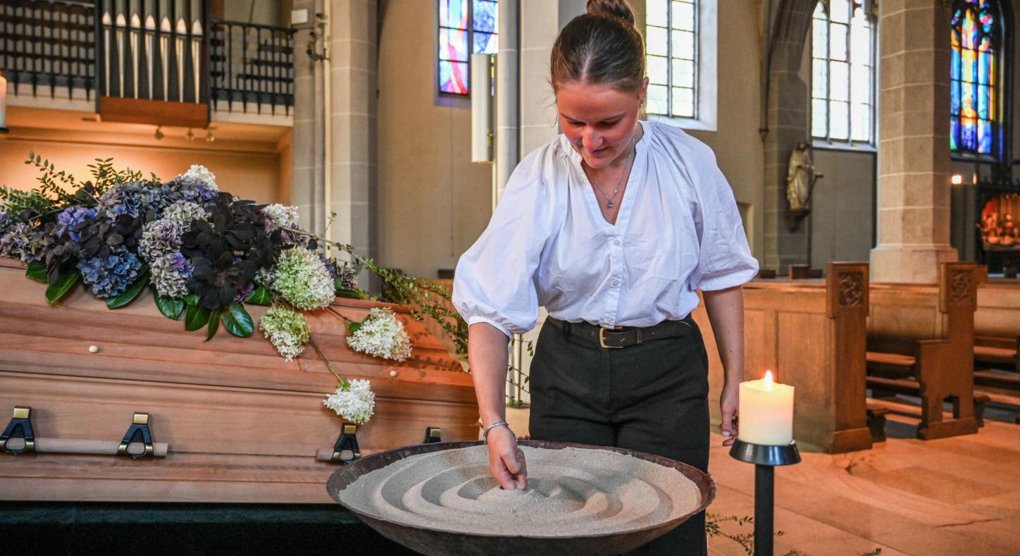 Eine Bestattering formt Sand in einer Schale, die vor einem geschmückten Sarg steht.