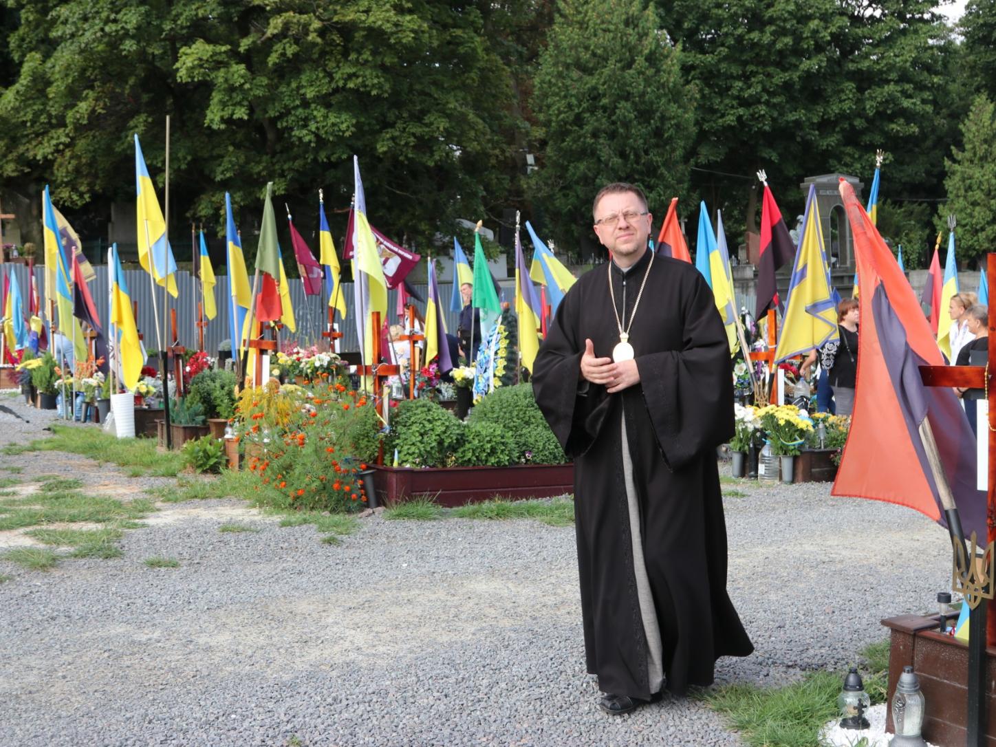 Mann mit Soutane in schwarz steht zwischen Fahnen auf einem Friedhof