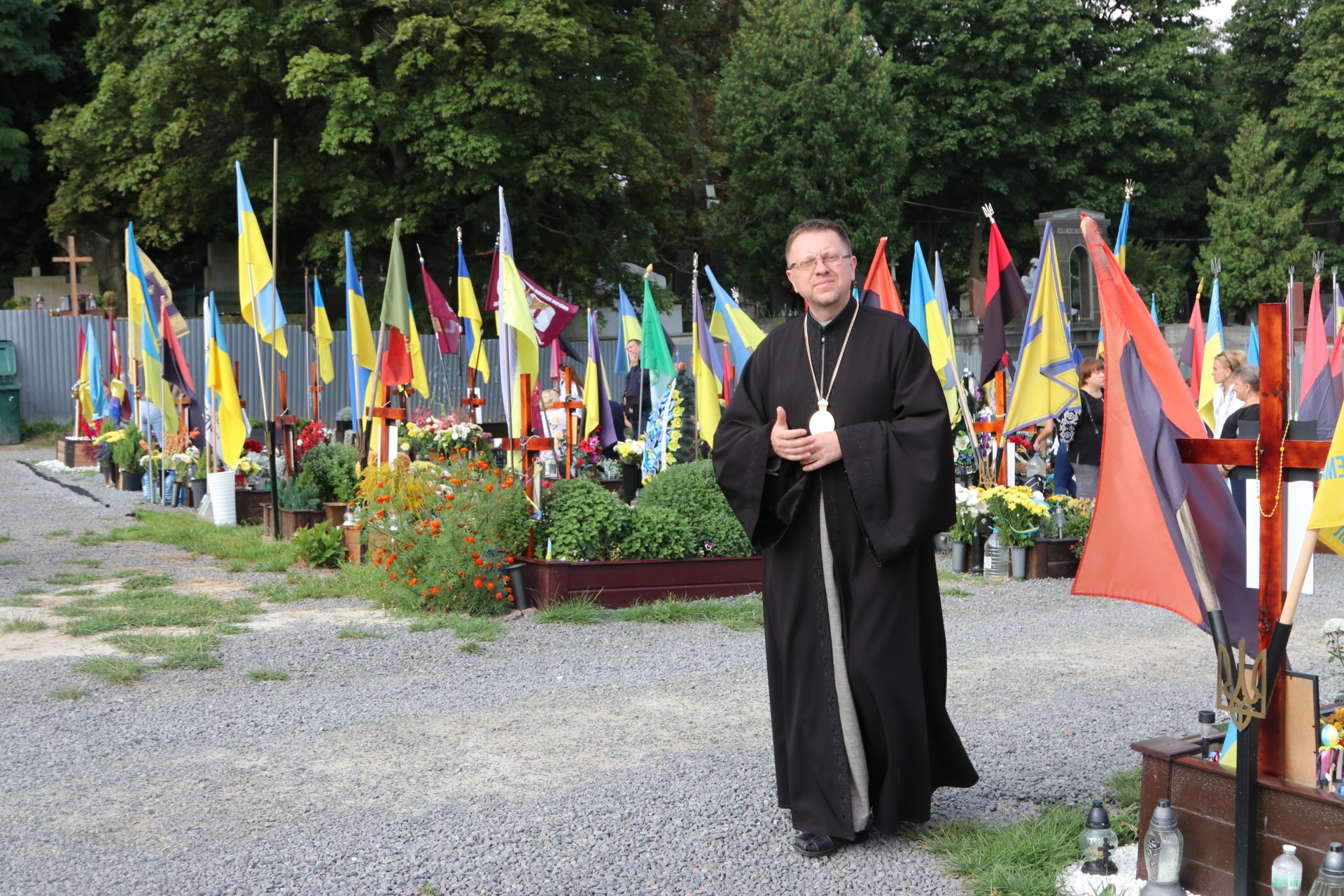 Mann mit Soutane in schwarz steht zwischen Fahnen auf einem Friedhof