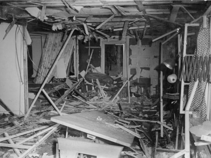 Das schwarz-weiße Foto zeigt einen völlig zerstörten Innenraum. In der Raummitte Reste eines Tischs, überall Holztrümmer, die Deckenverkleidung ist zerstört und hängt teilweise herunter, Fenster sind aus den Angeln gerissen. An der Wand auf der rechten Seite im Vordergrund eine Wandlampe mit Scherenarm, wie sie in Büroräumen verwendet werden.