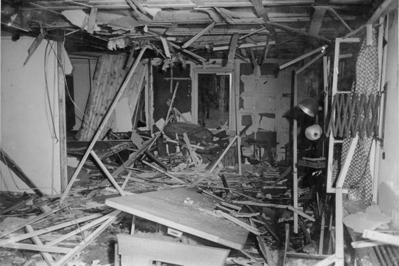 Das schwarz-weiße Foto zeigt einen völlig zerstörten Innenraum. In der Raummitte Reste eines Tischs, überall Holztrümmer, die Deckenverkleidung ist zerstört und hängt teilweise herunter, Fenster sind aus den Angeln gerissen. An der Wand auf der rechten Seite im Vordergrund eine Wandlampe mit Scherenarm, wie sie in Büroräumen verwendet werden.
