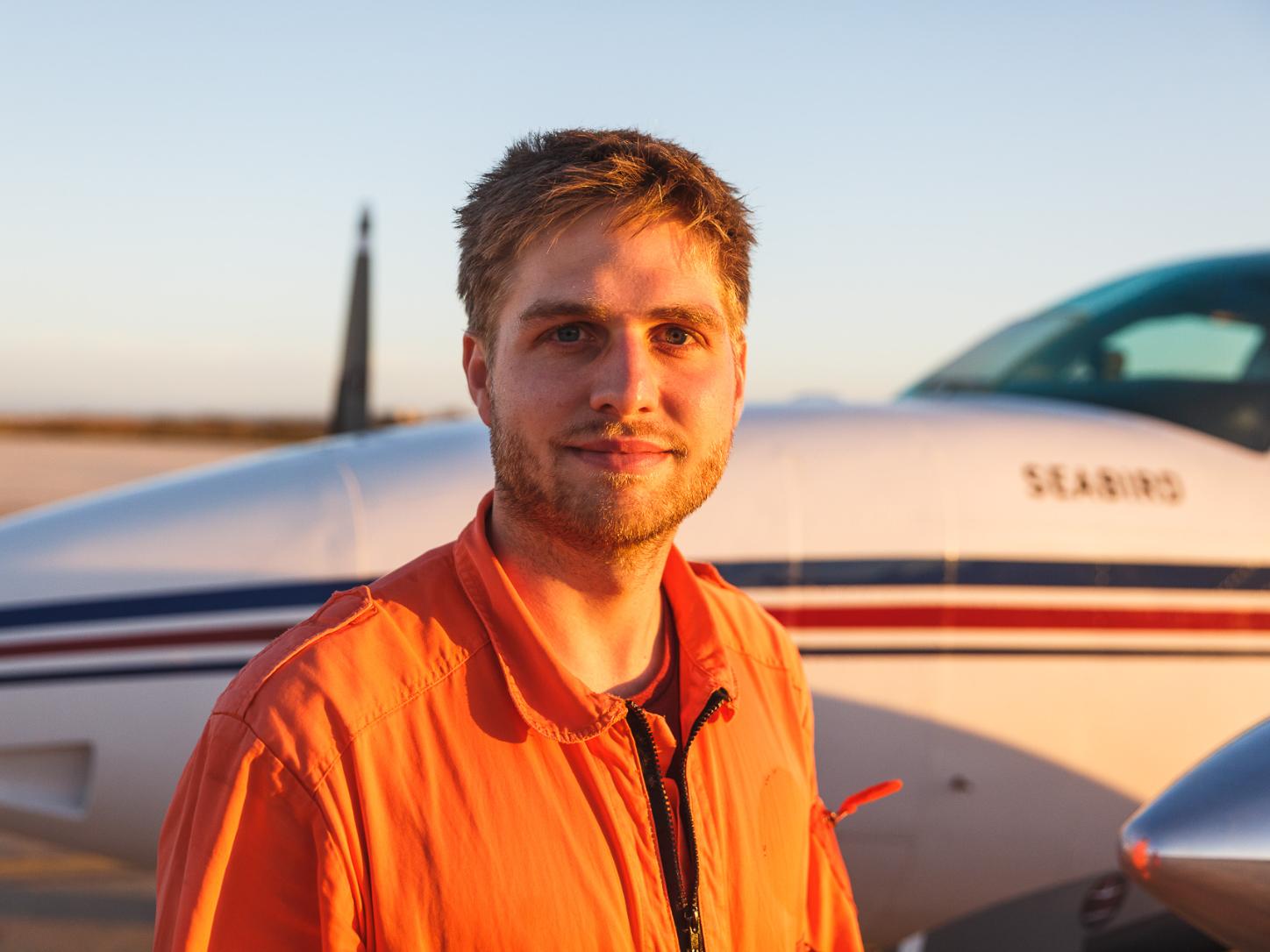 Mann in orangem Overall vor kleinem Flugzeug bei Sonnenuntergang.