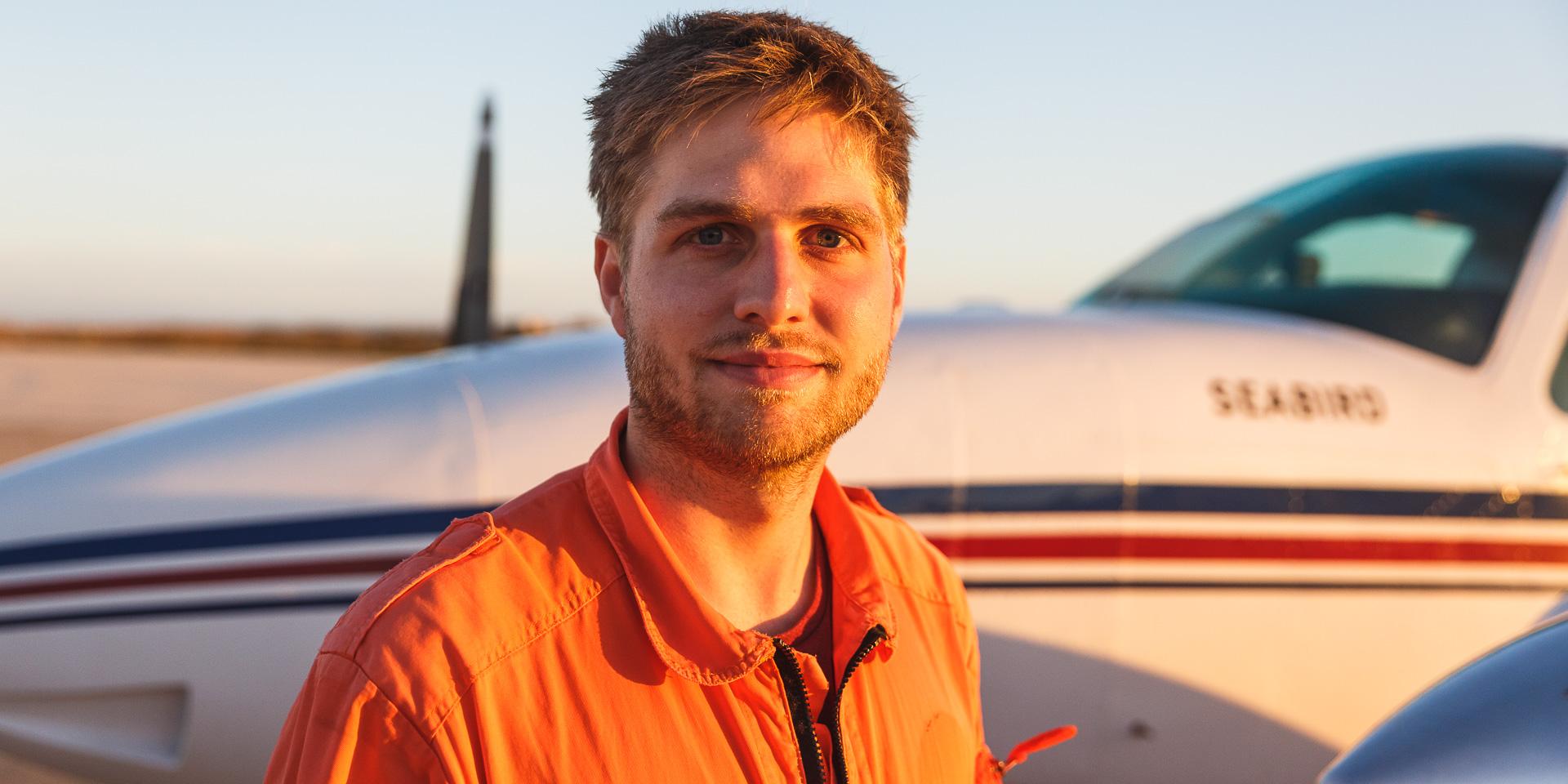 Mann in orangem Overall vor kleinem Flugzeug bei Sonnenuntergang.