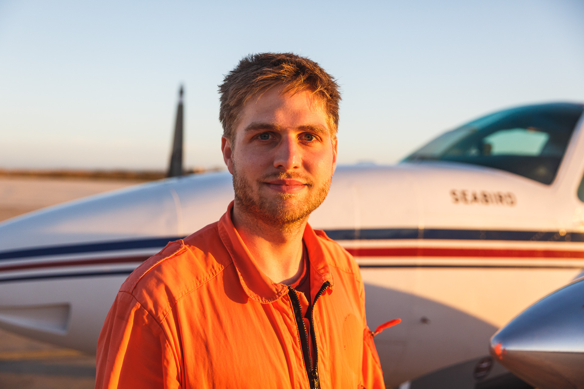 Mann in orangem Overall vor kleinem Flugzeug bei Sonnenuntergang.