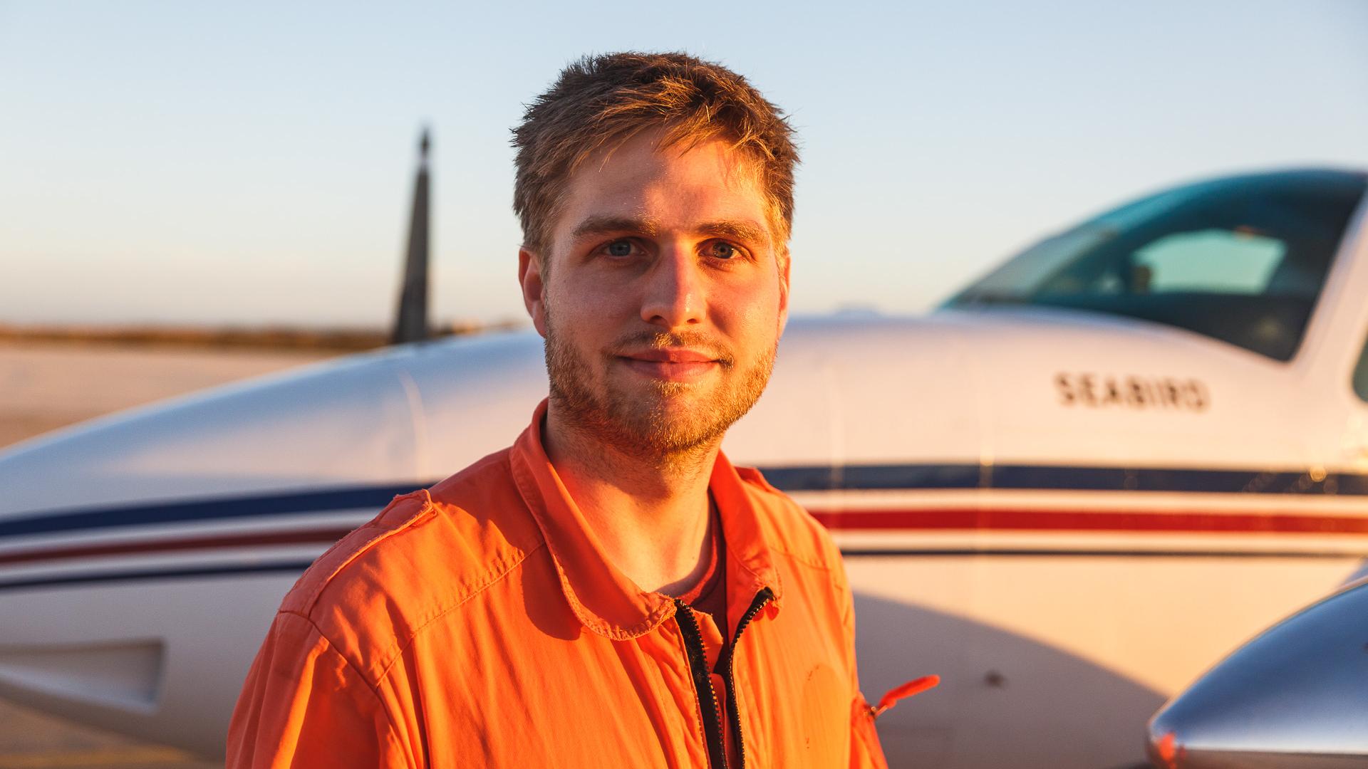 Mann in orangem Overall vor kleinem Flugzeug bei Sonnenuntergang.
