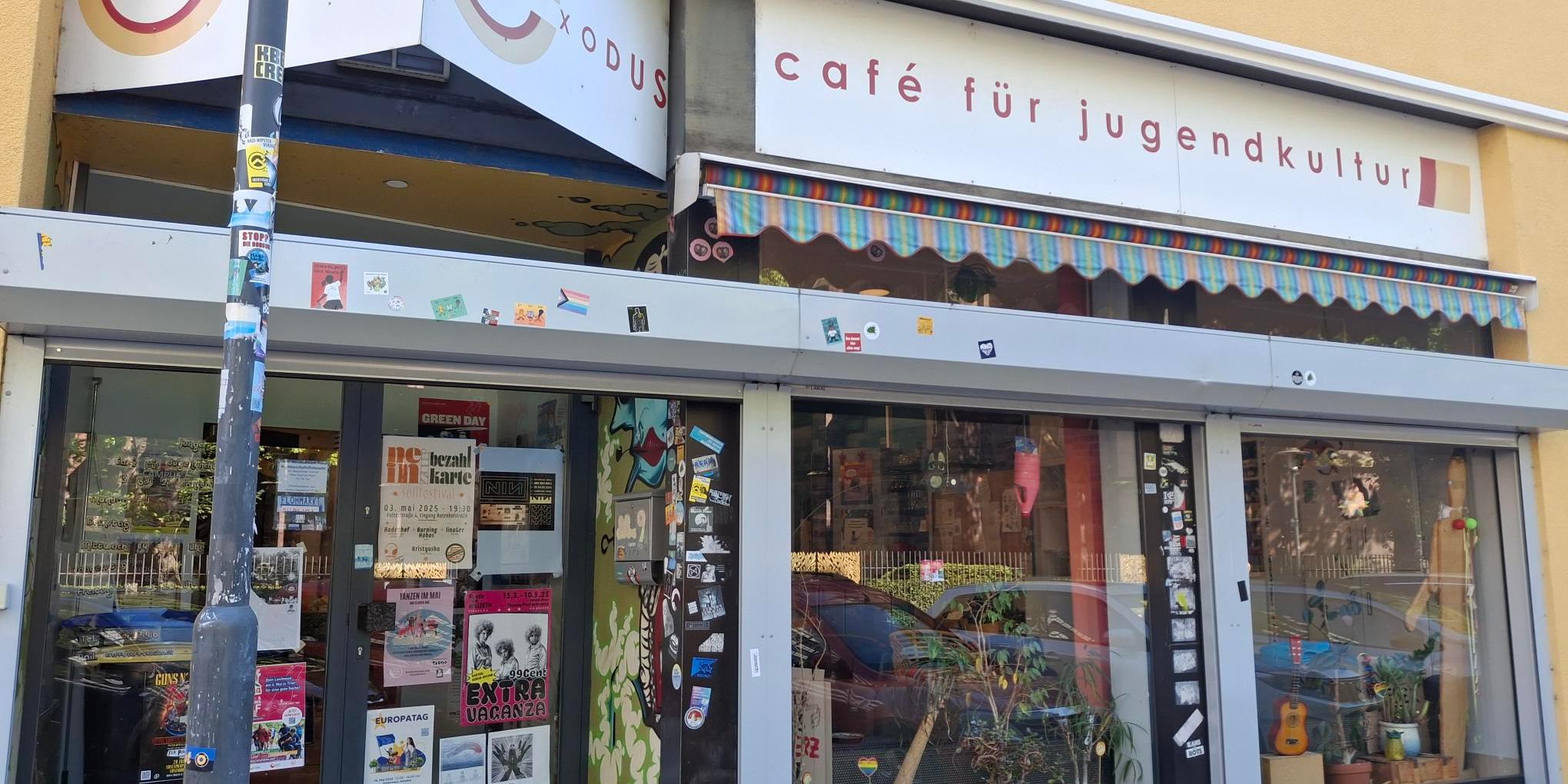 Eingang des ‚Café Exodus – Café für Jugendkultur‘ in einem gelben Gebäude. Über dem Eingang hängen zwei Schilder mit dem Schriftzug ‚Exodus‘ und ‚Café für Jugendkultur‘. Vor den großen Fenstern stehen viele Pflanzen, und die Glasflächen sind mit zahlreichen Stickern und Plakaten beklebt. Links befindet sich eine Glastür mit weiteren bunten Plakaten, die auf Veranstaltungen hinweisen. Ein Laternenpfahl steht am linken Bildrand und ist ebenfalls mit Stickern beklebt.