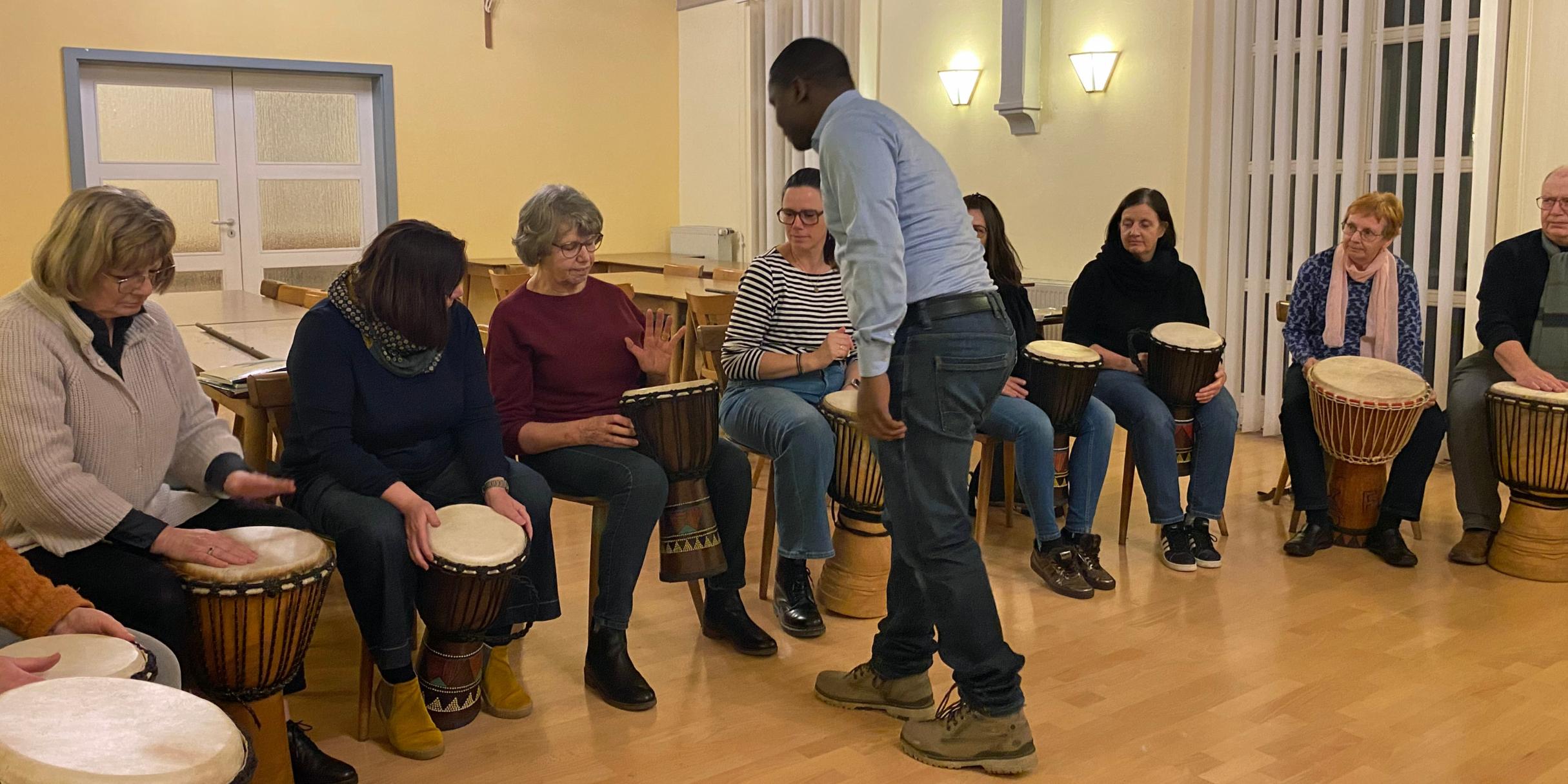 Eine Gruppe von Menschen sitzt in einem hell erleuchteten Raum in einem Halbkreis, jeweils mit einer Djembe‑Trommel vor sich. Ein stehender Anleiter in der Mitte gibt Hinweise oder führt etwas vor. Der Raum hat helle Holzböden, hohe Fenster mit Lamellen und warmgelbe Wände mit Wandlampen. Alle wirken konzentriert und in eine gemeinschaftliche Trommelübung vertieft.
