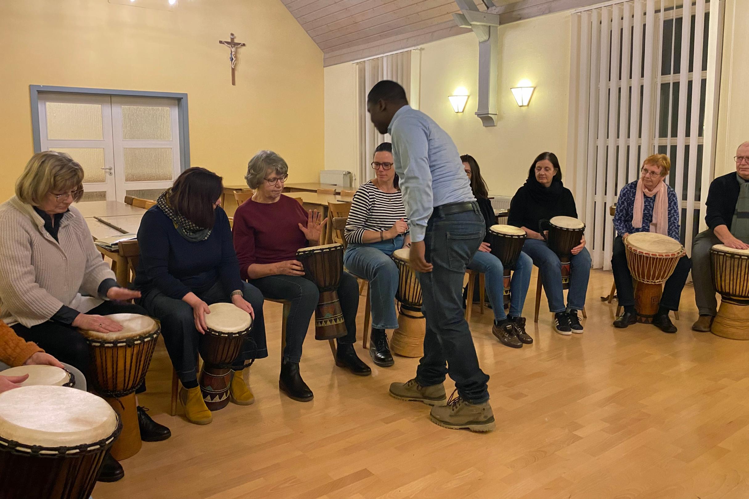 Eine Gruppe von Menschen sitzt in einem hell erleuchteten Raum in einem Halbkreis, jeweils mit einer Djembe‑Trommel vor sich. Ein stehender Anleiter in der Mitte gibt Hinweise oder führt etwas vor. Der Raum hat helle Holzböden, hohe Fenster mit Lamellen und warmgelbe Wände mit Wandlampen. Alle wirken konzentriert und in eine gemeinschaftliche Trommelübung vertieft.