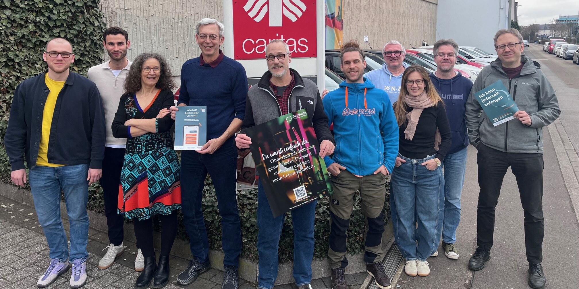 Eine Gruppe von unterschiedlichen Leuten steht draußen vor dem Caritas-Logo und schaut in die Kamera.