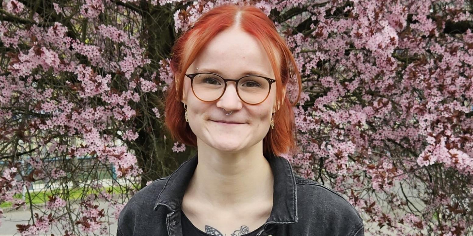 Eine junge Frau mit roten Haare und einer Brille schaut lächelnd in die Kamera.