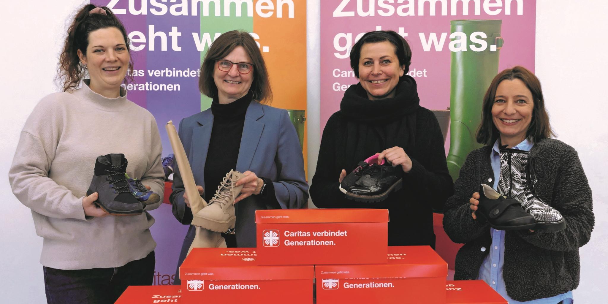 Vier Frauen lächeln in die Kamera und halten je einen Schuh hoch, vor ihnen stehen rote Schuhkartons.