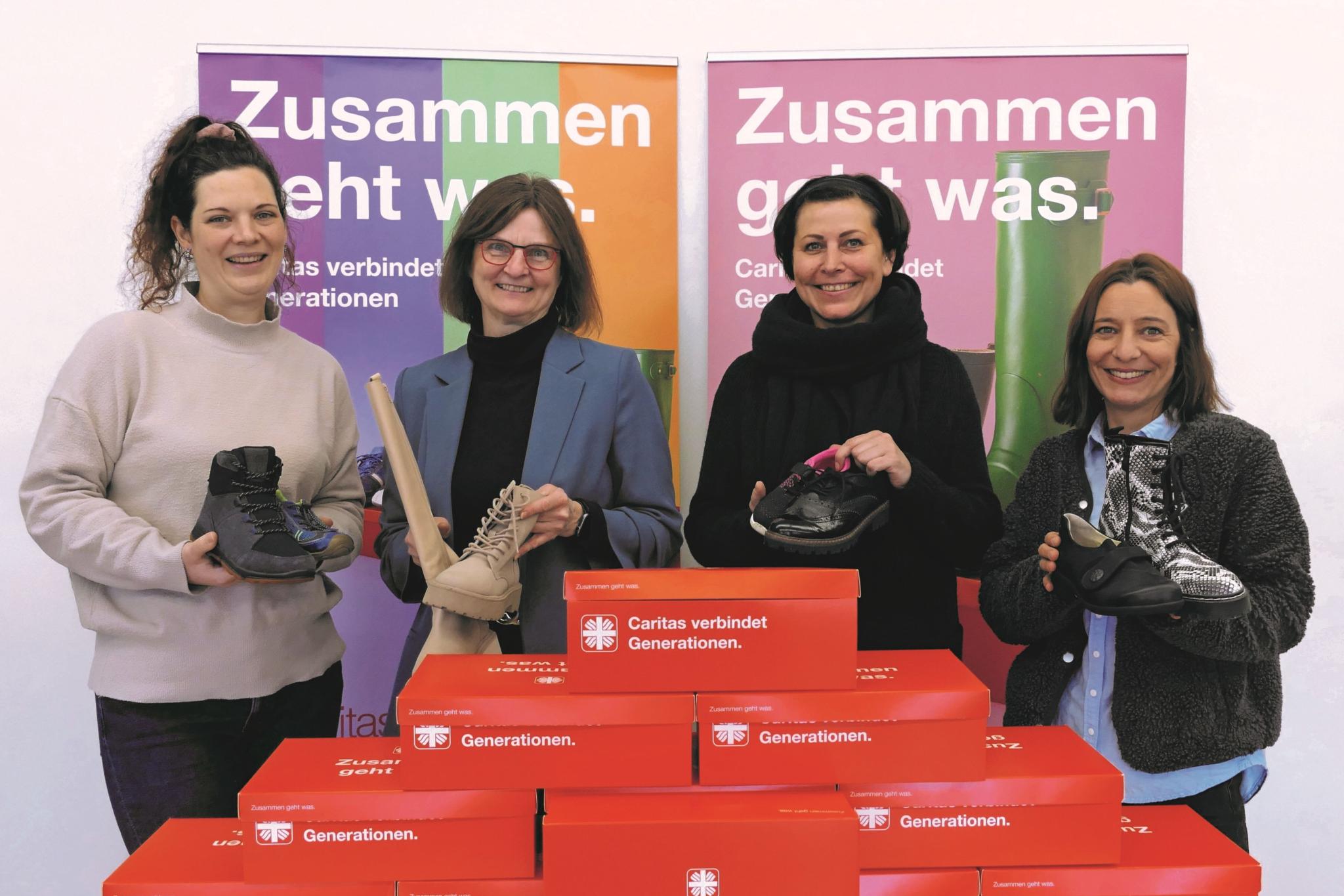 Vier Frauen lächeln in die Kamera und halten je einen Schuh hoch, vor ihnen stehen rote Schuhkartons.