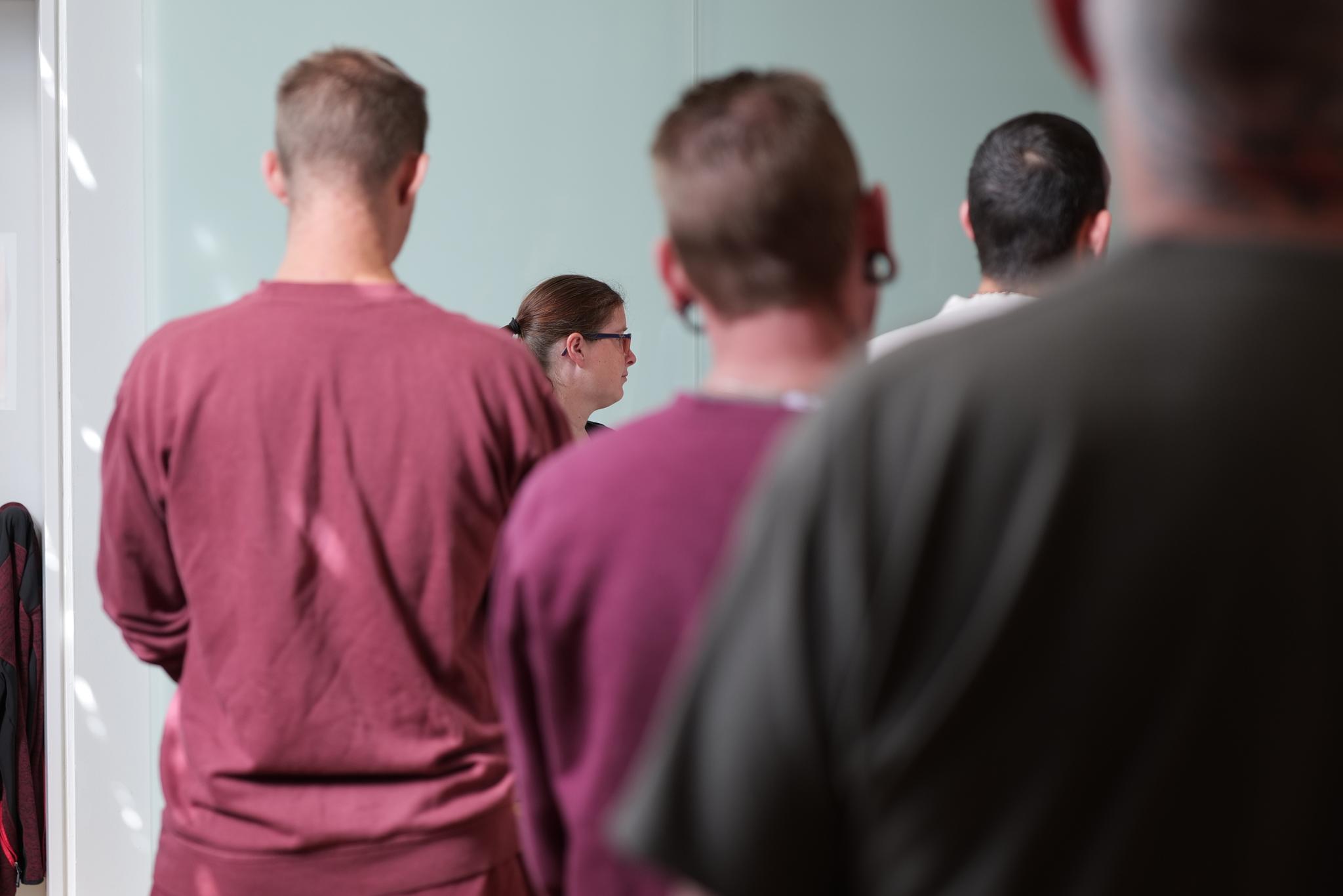Das Foto zeigt – teilweise unscharf - Oberkörper und Köpfe von vier Männern von hinten, zum Teil verdecken sie sich gegenseitig. Etwas im Hintergrund, zwischen zwei der Männer, der Kopf einer Frau im Profil; es handelt sich dabei um Clara Boor, die Hauptperson der Podcast-Folge. Über das Foto ist in dünnen weißen Großbuchstaben der Satz gelegt: „Clara Boor ist Organistin in der JVA“.