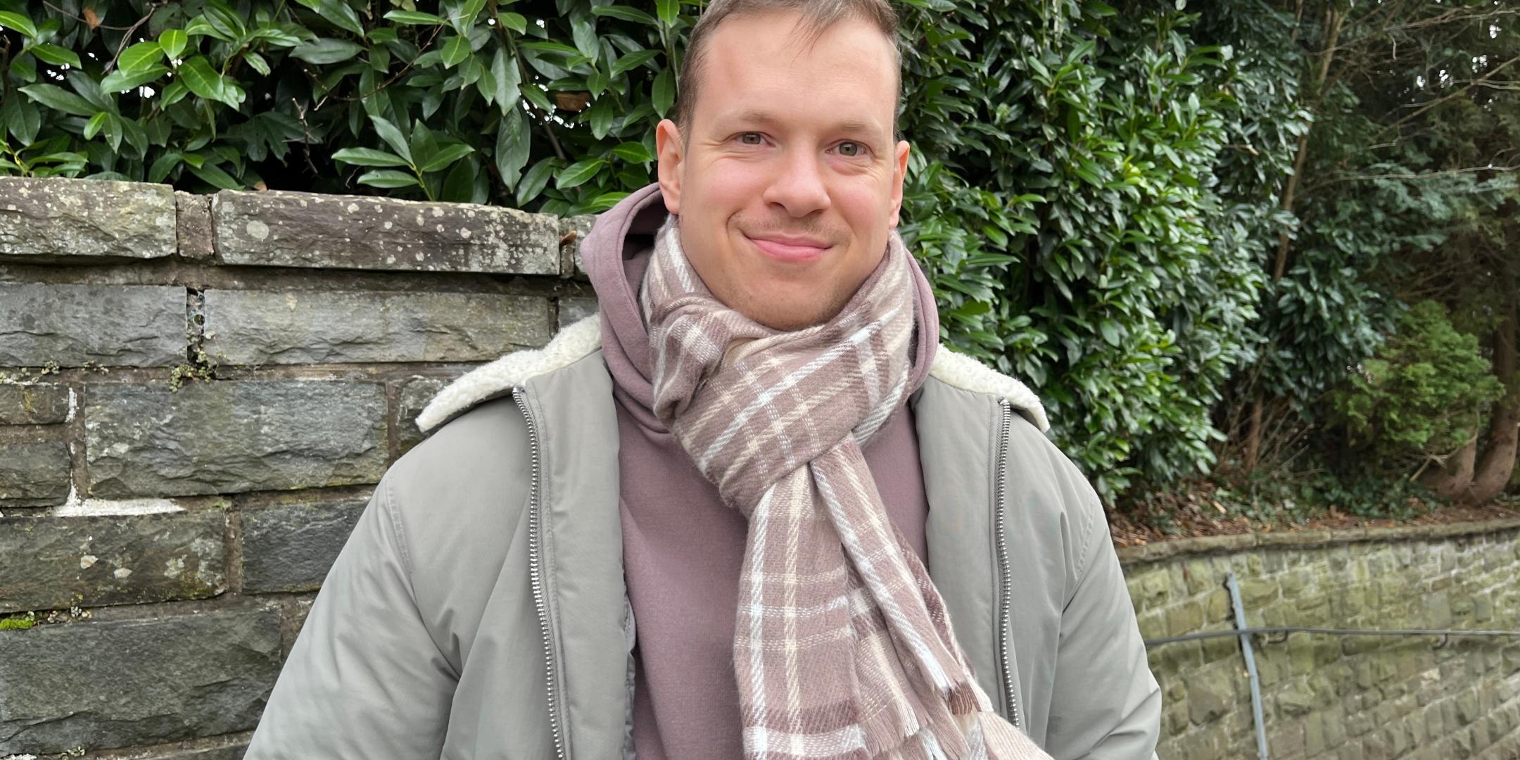 Ein junger Mann mit kurzem, hellbraunem Haar und freundlichem Lächeln steht vor einer Natursteinmauer mit dichten grünen Büschen im Hintergrund. Er trägt eine olivgrüne Winterjacke mit heller Kapuze, einen braunen Kapuzenpullover und einen beigefarbenen karierten Schal, der um seinen Hals gewickelt ist. Seine Hände sind in den Jackentaschen, und er blickt direkt in die Kamera. (Alt-Text erstellt mit Unterstützung von ChatGPT)