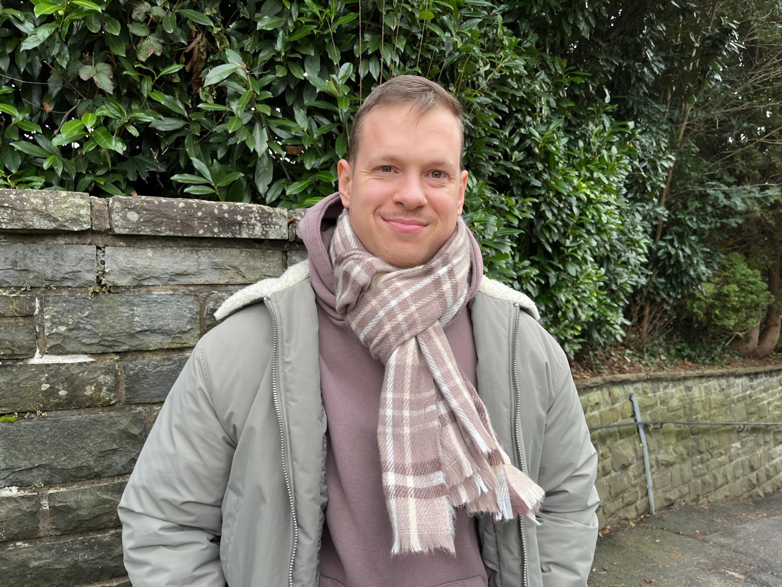 Ein junger Mann mit kurzem, hellbraunem Haar und freundlichem Lächeln steht vor einer Natursteinmauer mit dichten grünen Büschen im Hintergrund. Er trägt eine olivgrüne Winterjacke mit heller Kapuze, einen braunen Kapuzenpullover und einen beigefarbenen karierten Schal, der um seinen Hals gewickelt ist. Seine Hände sind in den Jackentaschen, und er blickt direkt in die Kamera. (Alt-Text erstellt mit Unterstützung von ChatGPT)