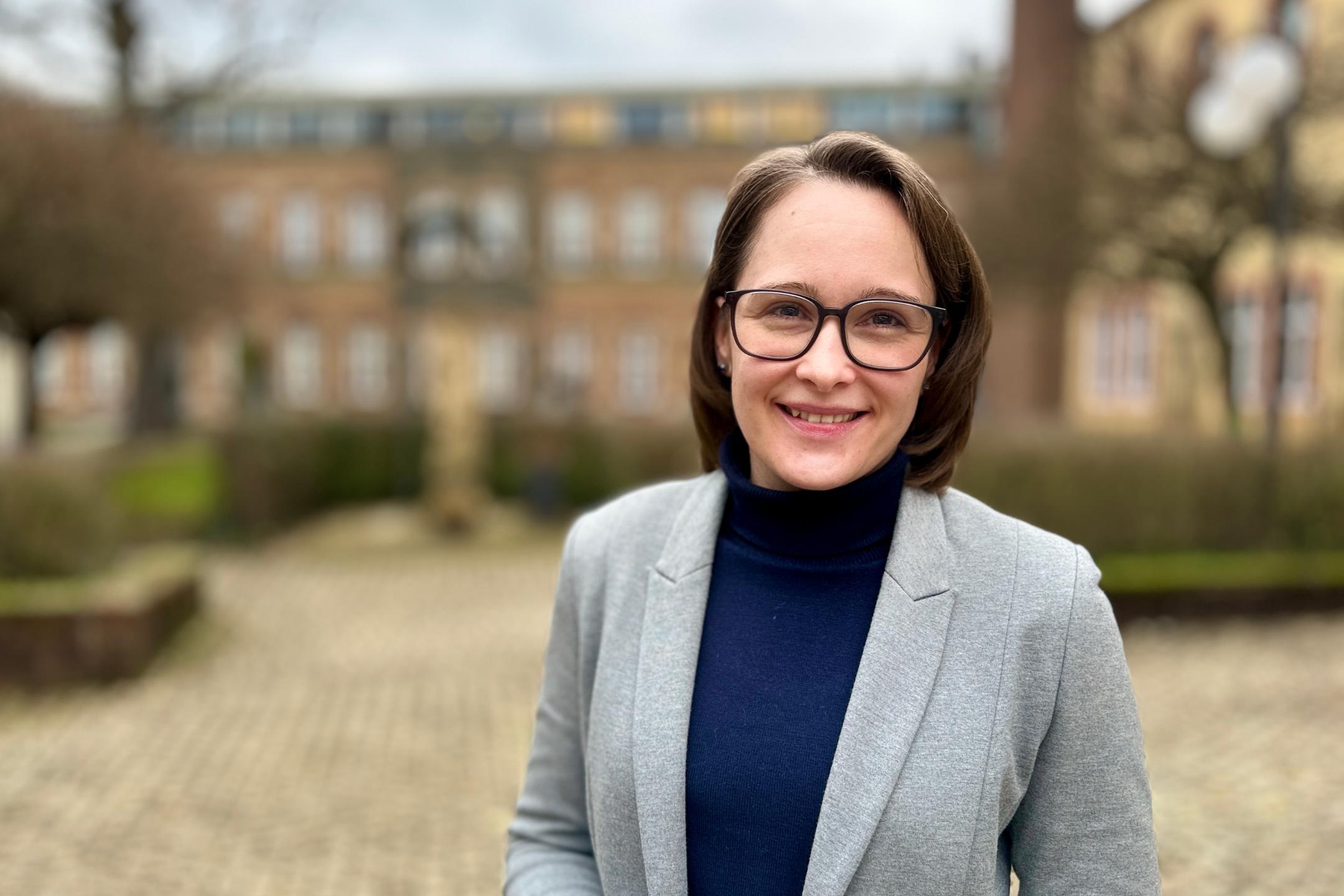 Eine Frau mit schulterlangem braunen Haar und Brille steht lächelnd im Freien vor einem historischen Backsteingebäude. Sie trägt einen grauen Blazer über einem dunkelblauen Rollkragenpullover. Der Hintergrund ist leicht unscharf, mit einem gepflasterten Weg, Hecken und Bäumen.