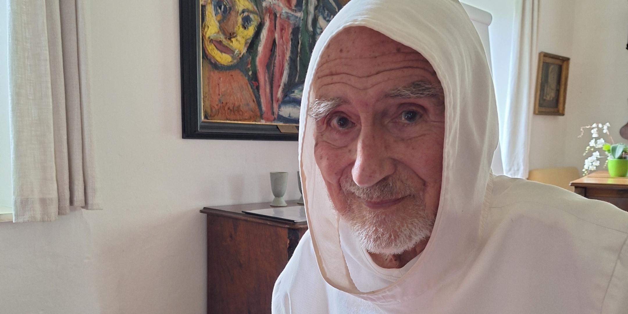 uf dem Foto sitzt der Benediktinermönch David Steindl-Rast in einem Raum an einem Tisch mit roter Decke. Er trägt ein weißes Ordensgewand mit Kapuze. Sein altes, schmales Gesicht mit einem kurzen weißen Bart ist dem Betrachter zugewandt, die Augen wirken sehr aufmerksam, sein Lächeln ist sehr freundlich.