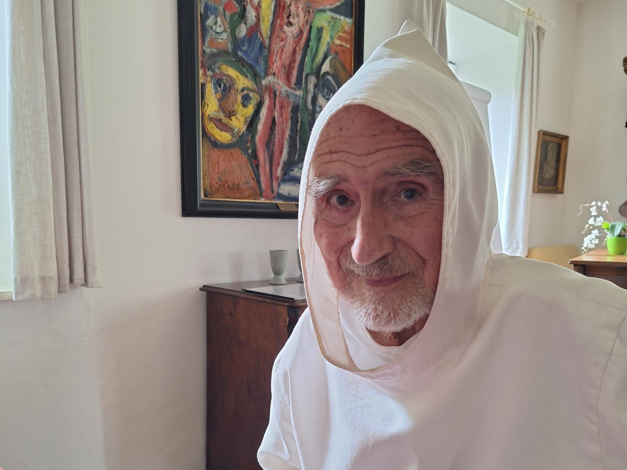 uf dem Foto sitzt der Benediktinermönch David Steindl-Rast in einem Raum an einem Tisch mit roter Decke. Er trägt ein weißes Ordensgewand mit Kapuze. Sein altes, schmales Gesicht mit einem kurzen weißen Bart ist dem Betrachter zugewandt, die Augen wirken sehr aufmerksam, sein Lächeln ist sehr freundlich.