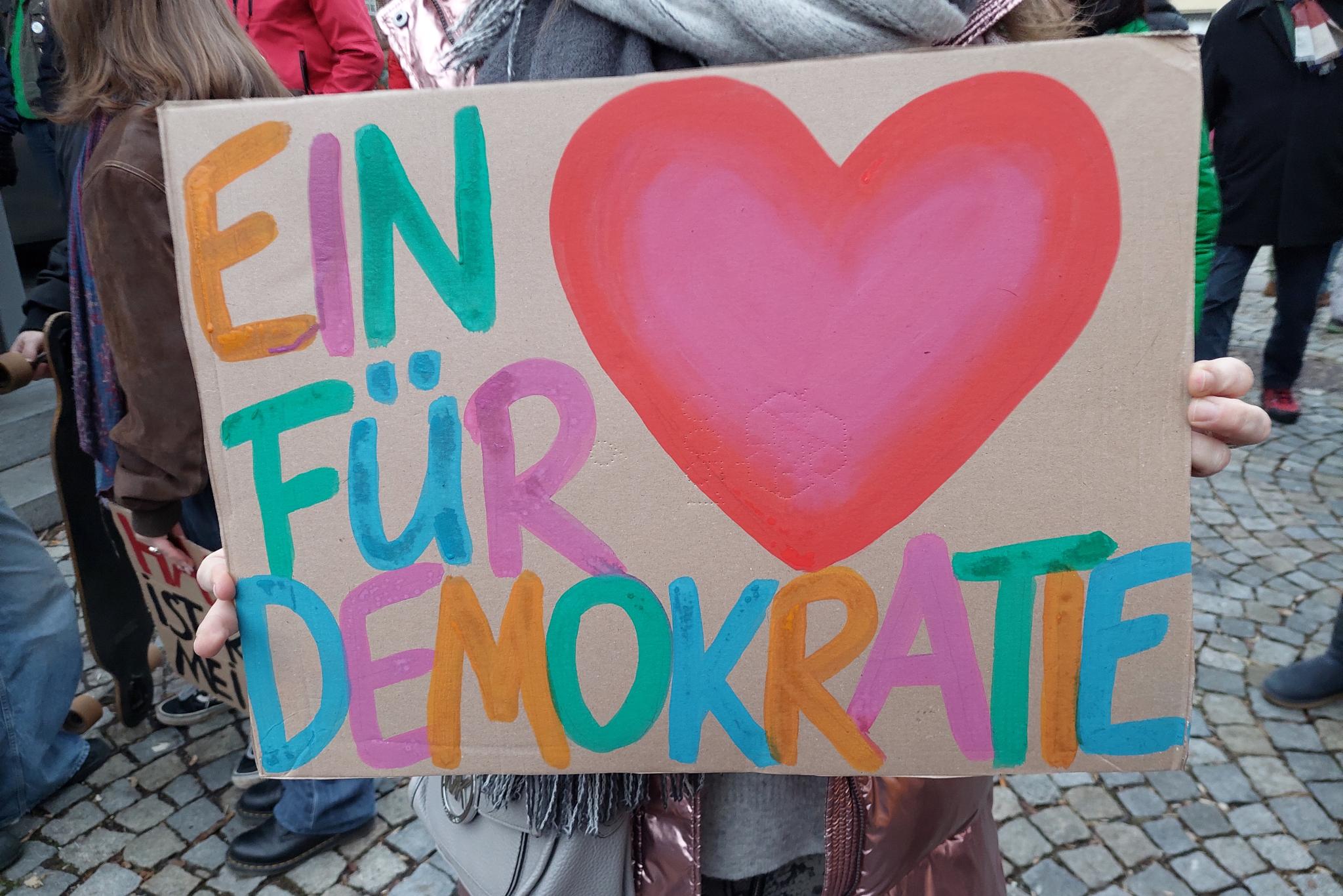 Eine Person hält ein Plakat in die Kamera. Darauf zu sehen ist ein der Schriftzug 'Ein Herz für Demokratie'.