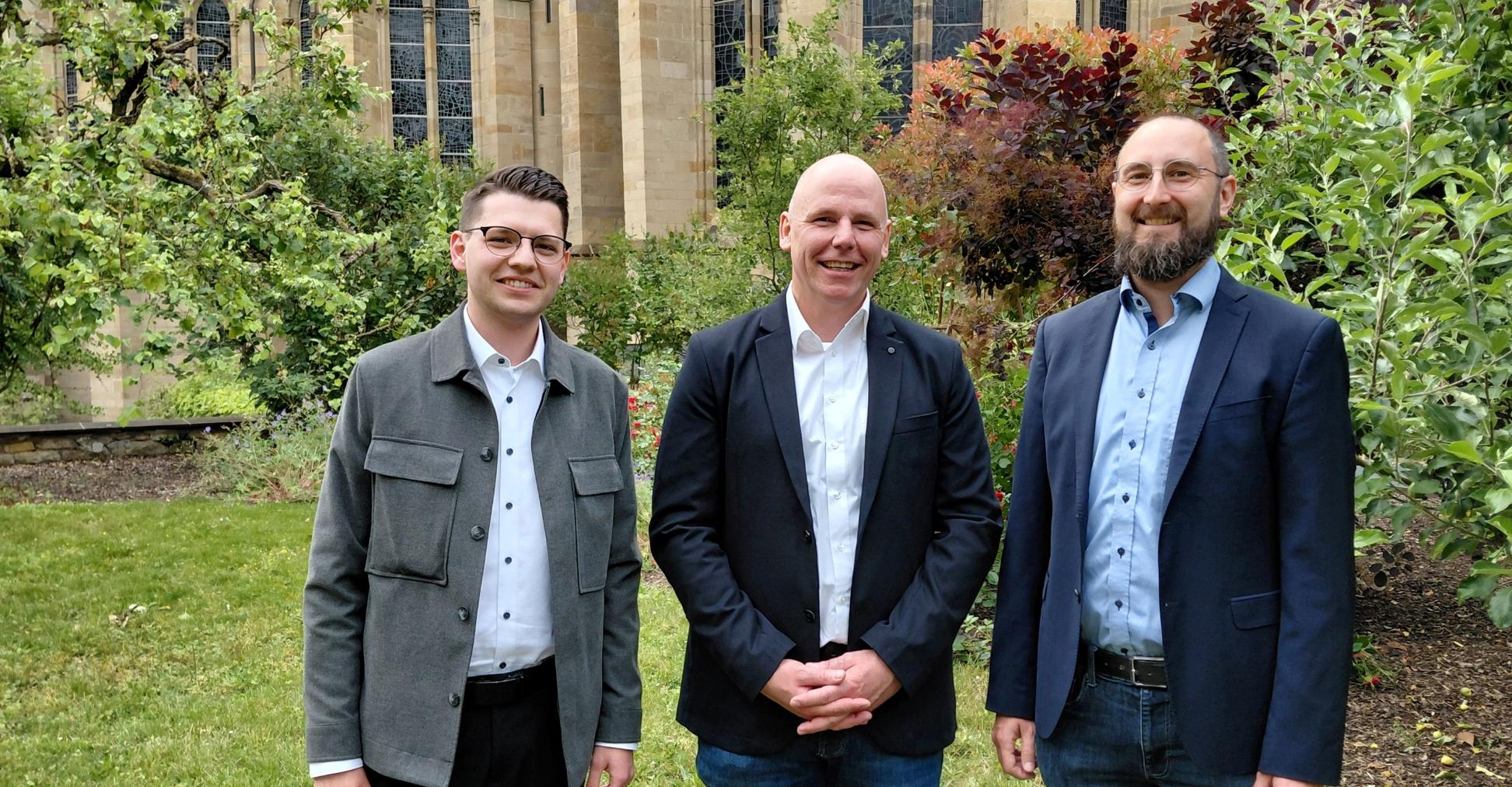 Drei Männer stehen lächelnd im Grünen; Im Hintergrund die Liebfrauenkirche