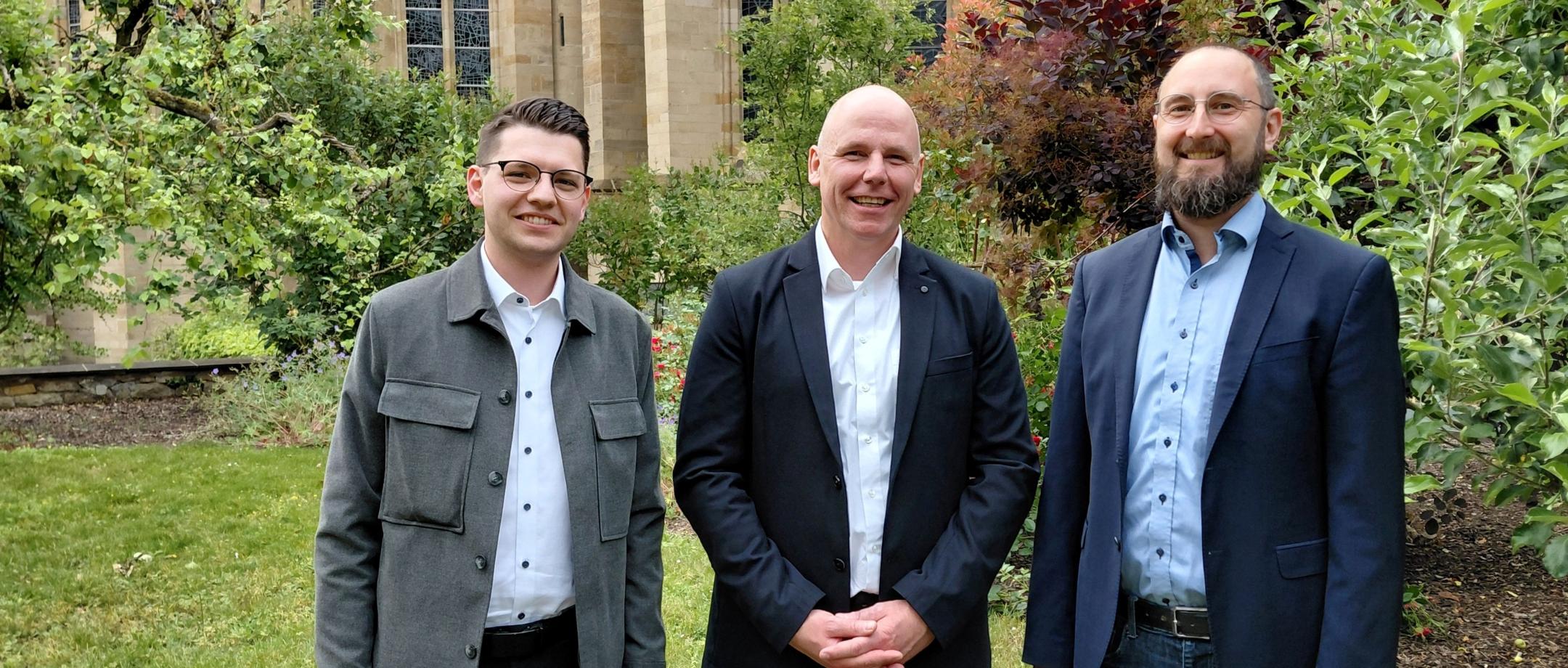 Drei Männer stehen lächelnd im Grünen; Im Hintergrund die Liebfrauenkirche