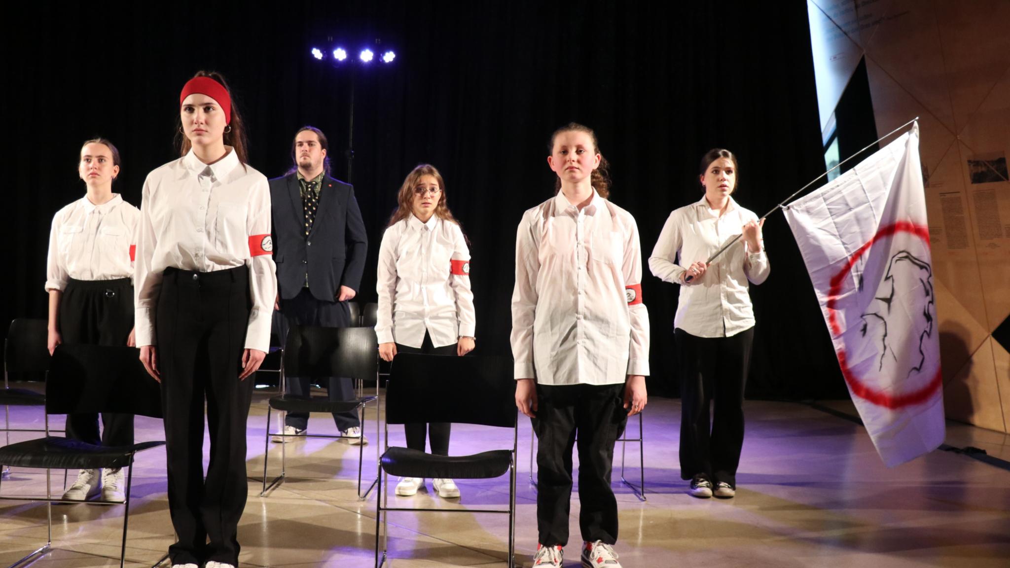 Die Gruppendynamik macht sich auch Eine Schülergruppe mit Lehrer steht 'stramm' neben ihren Stühlen. Die Schüler tragen alle weiße Blusen, schwarze Hosen und ein rotes Armband mit Wellensymbol.