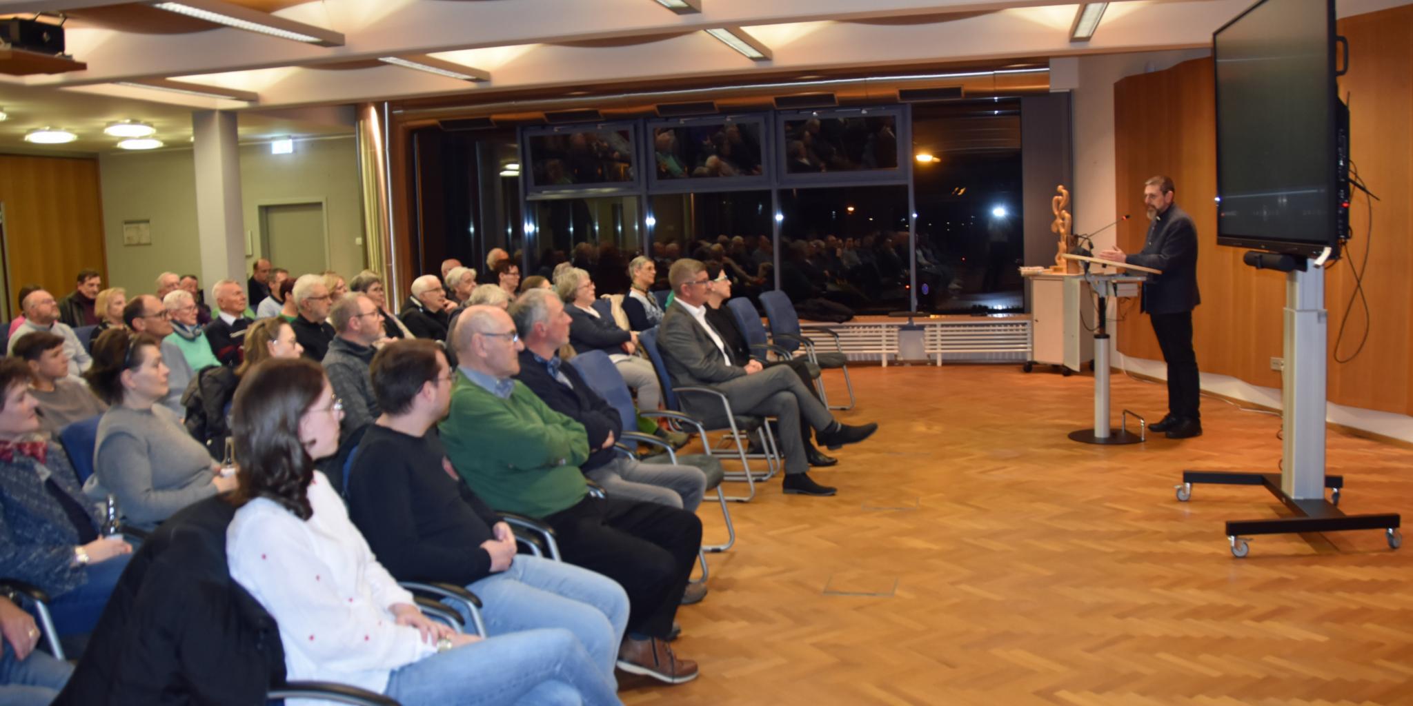 Etwa 80 Menschen sitzen auf Stühlen und hören den Worten des Dekans zu. Neben dem Dekan steht ein großes Fernsehgerät