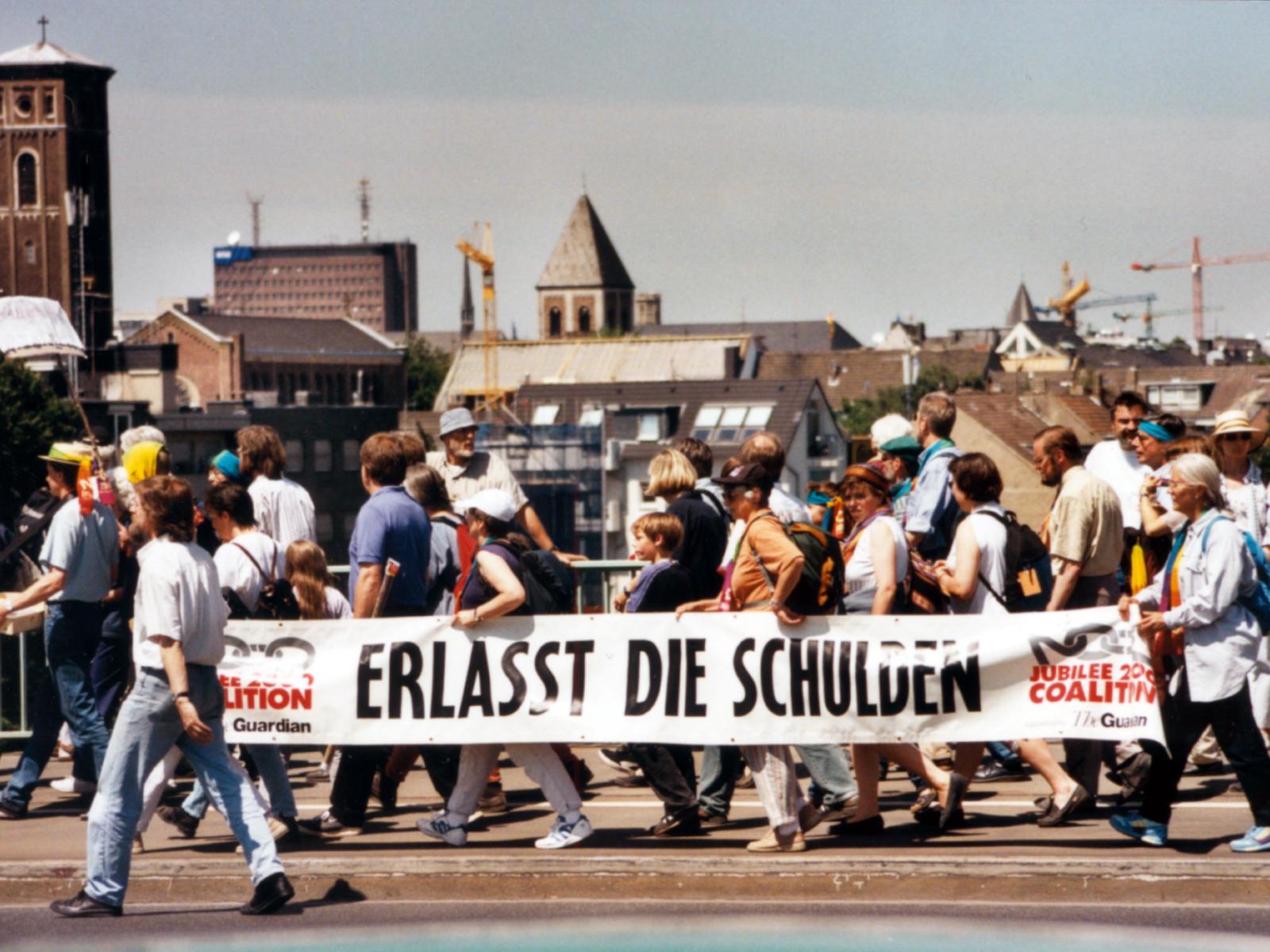 Menschen bei einer Protestaktion tragen ein großes Banner mit der Aufschrift 'Erlasst die Schulden'