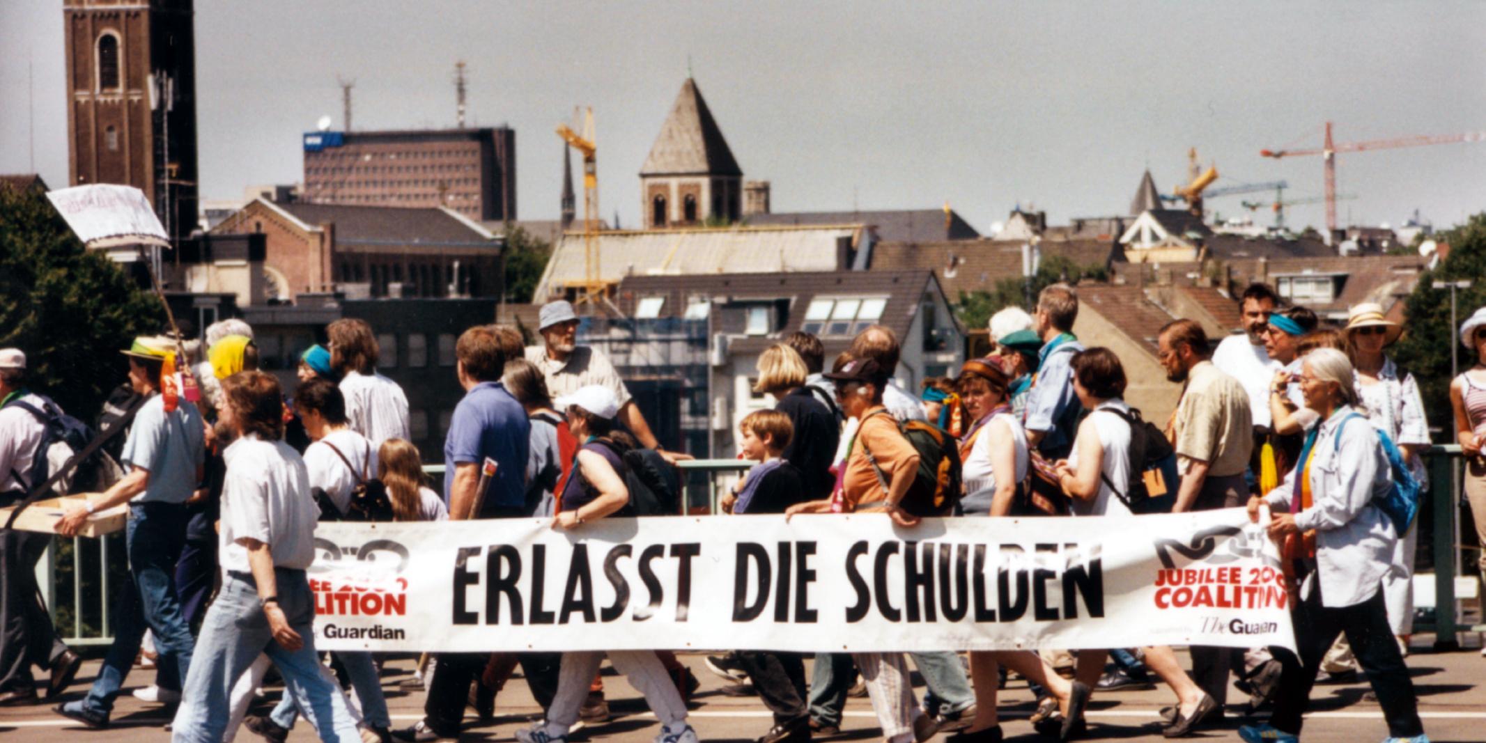Menschen bei einer Protestaktion tragen ein großes Banner mit der Aufschrift 'Erlasst die Schulden'