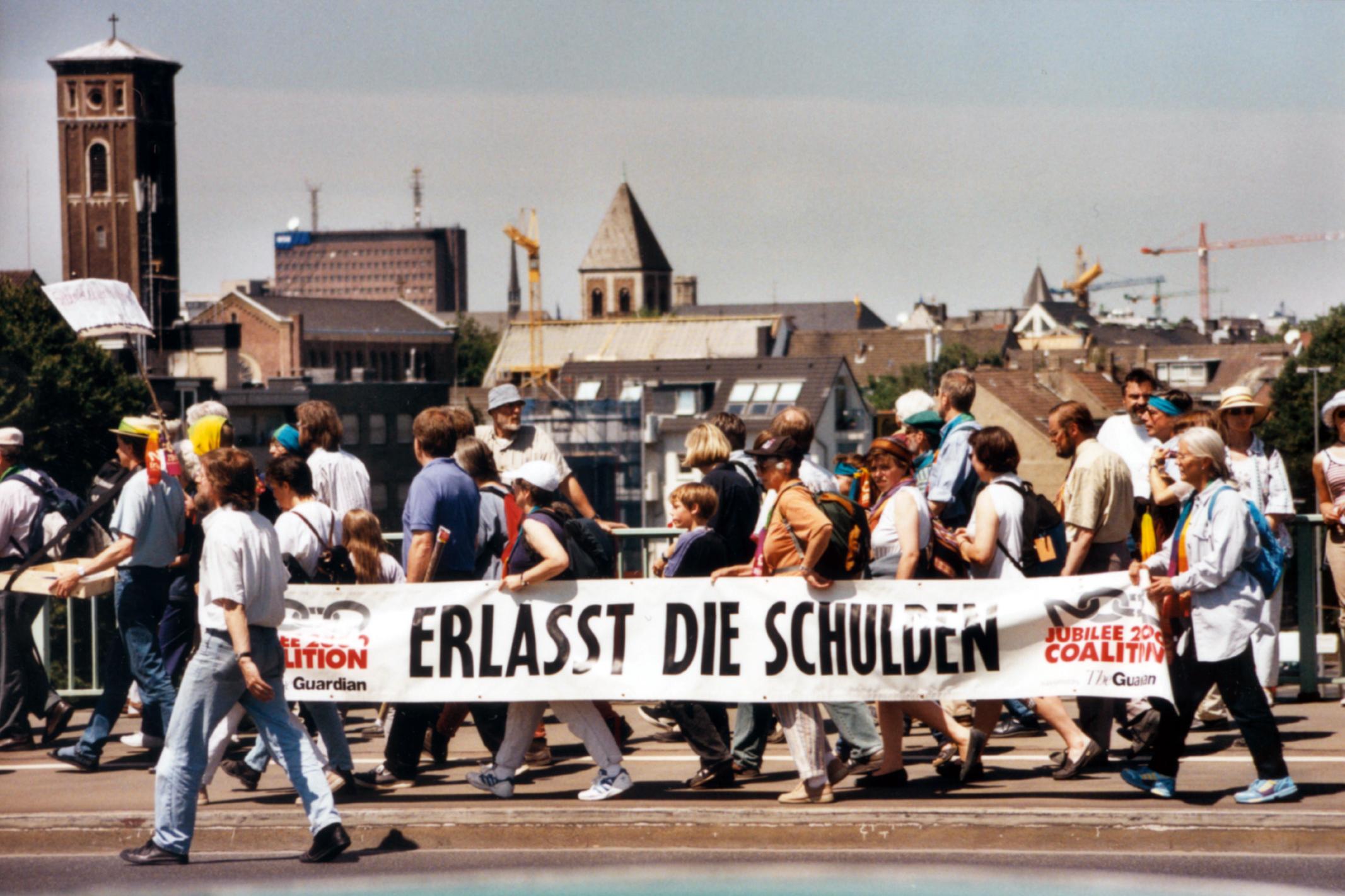 Menschen bei einer Protestaktion tragen ein großes Banner mit der Aufschrift 'Erlasst die Schulden'