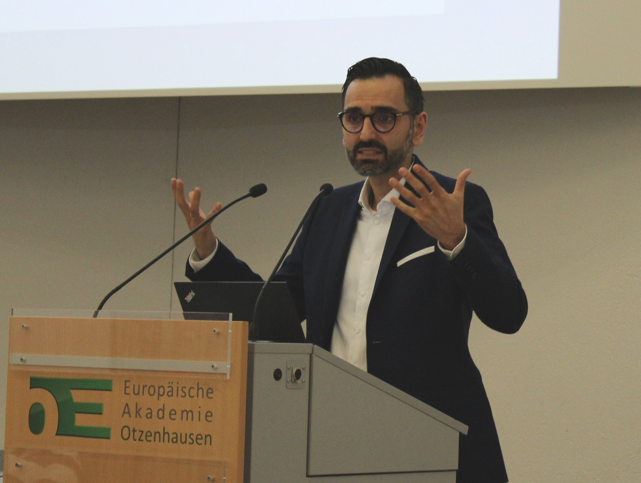 Eine Person steht hinter einem Rednerpult der Europäischen Akademie Otzenhausen und hält einen Vortrag. Die Person trägt einen dunklen Anzug und gestikuliert mit beiden Händen. Vor ihr stehen zwei Mikrofone und ein Laptop. Im Hintergrund ist eine helle Präsentationsfläche zu sehen.