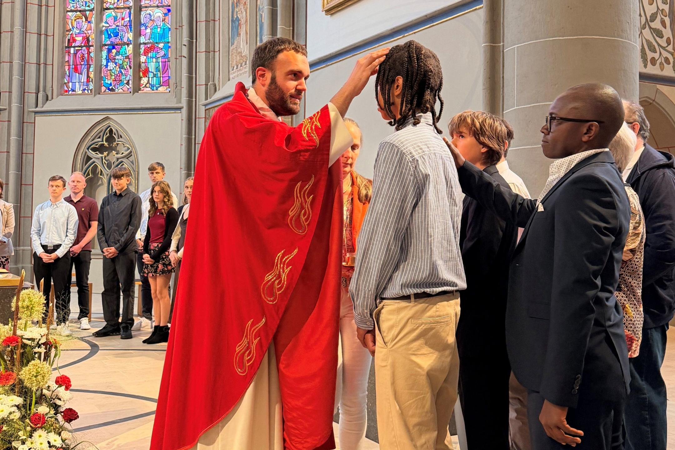 Ein Priester in einem roten Gewand mal mit seinem Finger ein Kreuzzeichen aus Öl auf die Stirn eines Jugendlichen. Hinter ihm steht sein Pate und legt seine Hand auf die Schulter des Jungen.