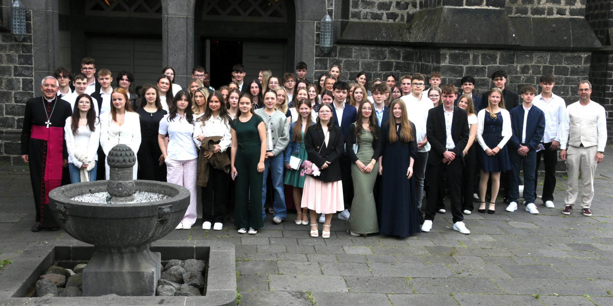 Gruppenfoto mit vielen jungen Menschen vor einer Kirche mit dunklen Steinen.