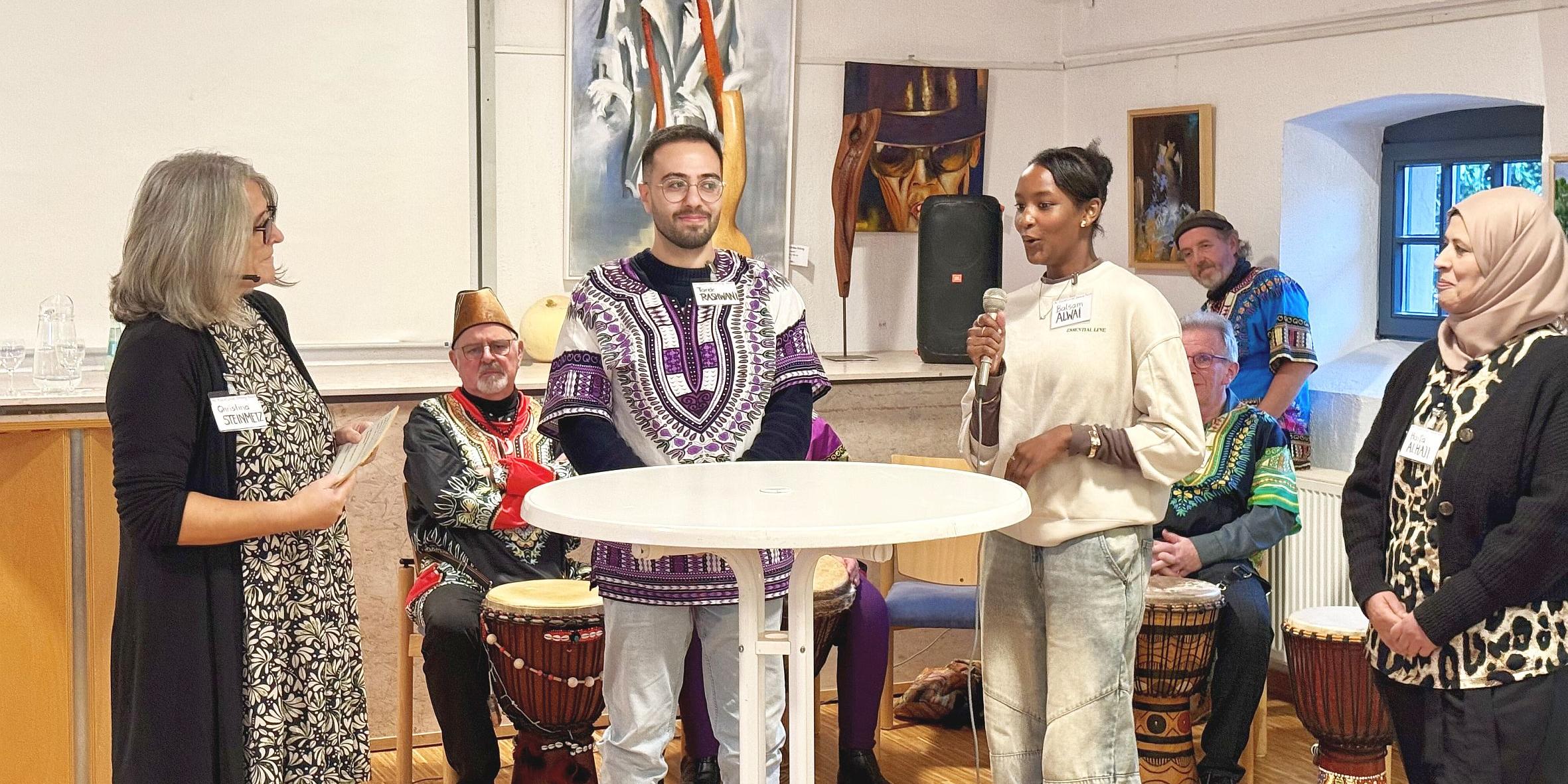 Vier Personen an einem Stehtisch, eine davon spricht in ein Mikrofon. Im Hintergrund sitzen Trommler in Tracht.