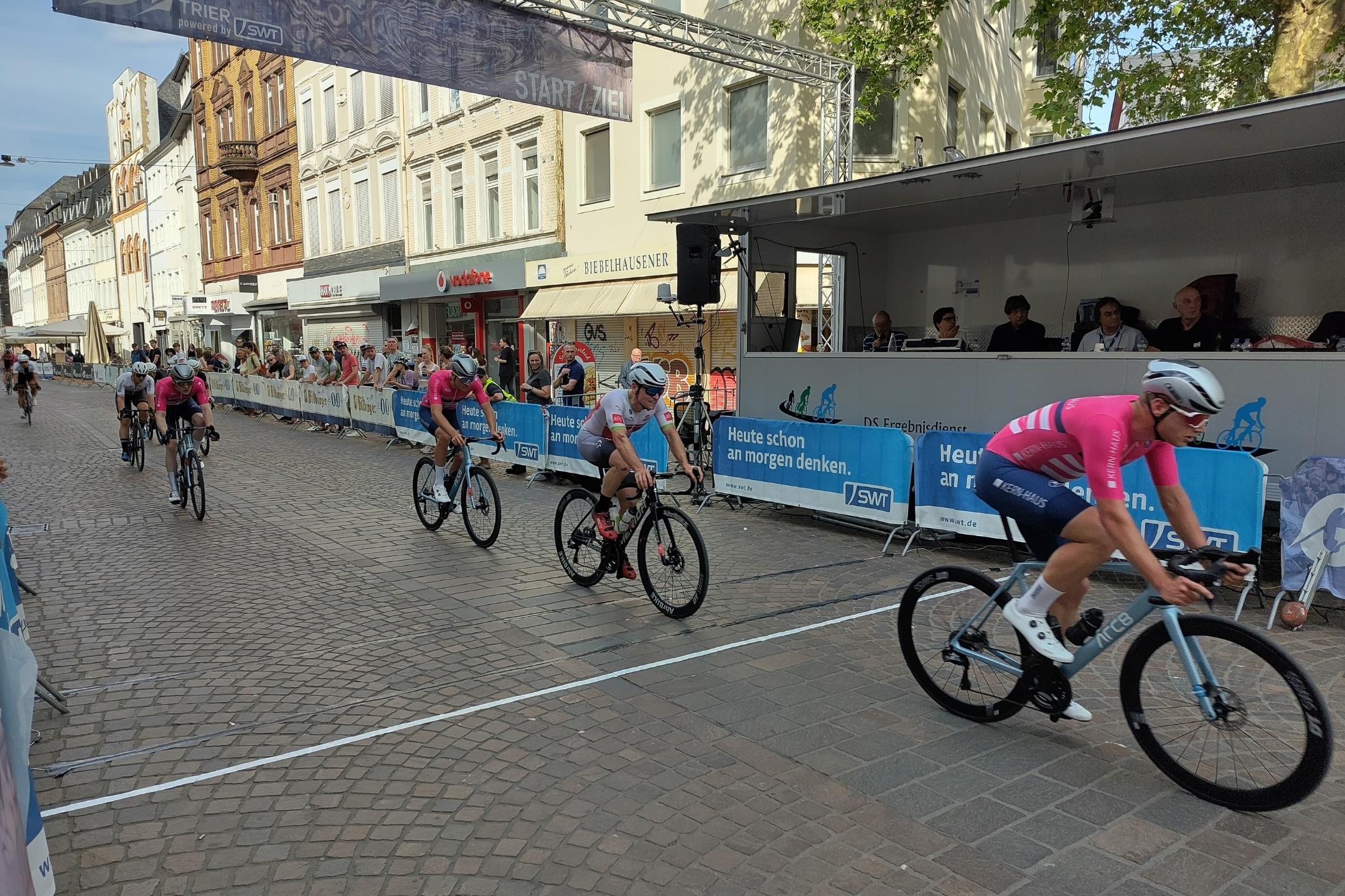 Foto eines Radrennens. Auf einer abgesperrten Innenstadtstraße (es handelt sich um einen Abschnitt der Trierer Fußgängerzone) fährt ein halbes Dutzend Radsportler von links nach rechts durch Bild, weiter hinten folgen weitere Fahrer. Hinter den Absperrungen am Rand stehen Zuschauer*innen.