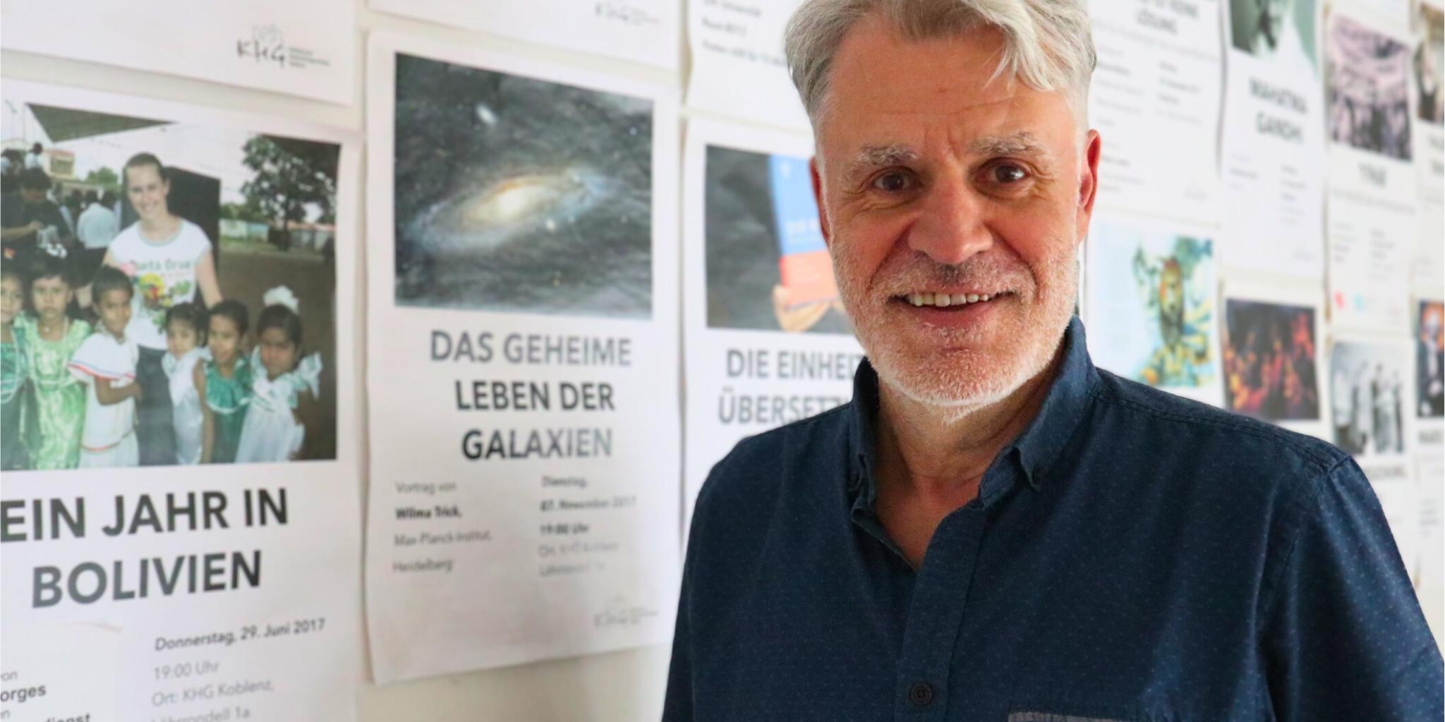 Auf dem Foto steht Guido Groß vor einer Pinnwand mit Veranstaltungsankündigungen der Katholischen Hochschulgemeinde Koblenz. Zu sehen sind sein Oberkörper und der Kopf. Er trägt ein kurzärmliges blaues Hemd, hat graue Haare und einen grauen Drei-Tage-Bart und schaut lächelnd in die Kamera.