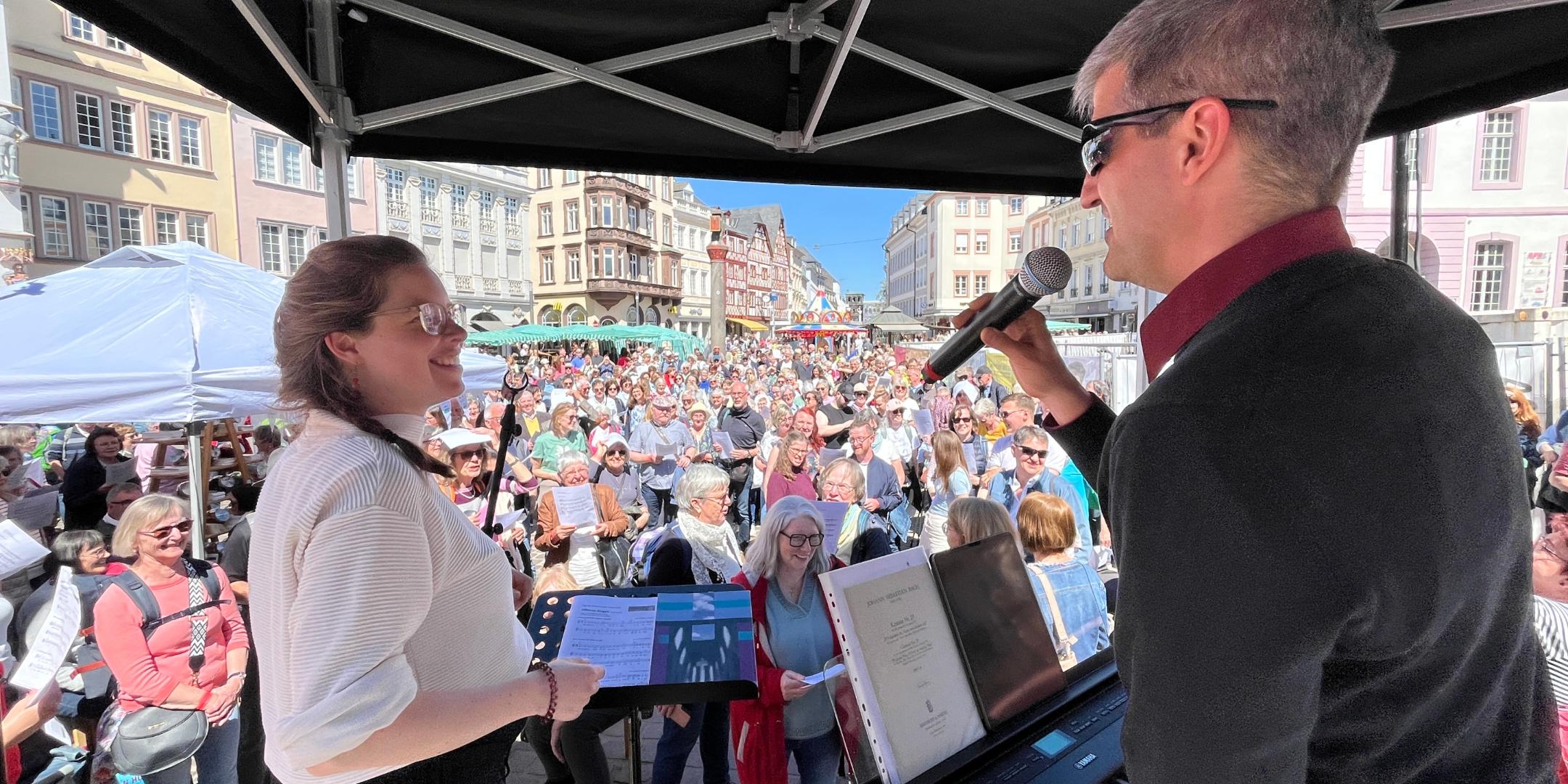 Eine Frau und ein Mann haben Mikrofone in der Hand, der Mann spielt Keyboard und sie stehen auf einer Bühne vor der sehr viele Leute auf einem Platz stehen.