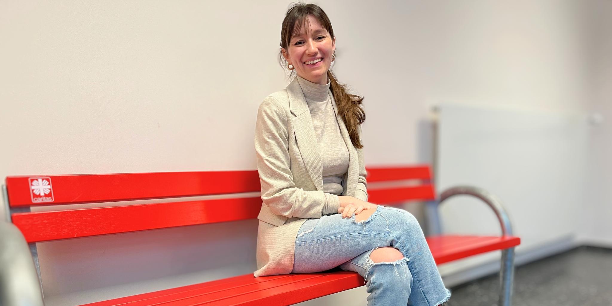 Auf dem Foto sitzt Hannah Matthews auf einer roten Bank mit einem kleinen Caritas-Logo. Sie trägt Jeans, einen hellen, beigefarbenen Pulli und eine Jacke in fast gleicher Farbe. Der Kopf ist nach rechts zur Kamera gewendet, die langen braunen Haare fallen in einem Pferdeschwanz über die linke Schulter, sie lächelt.