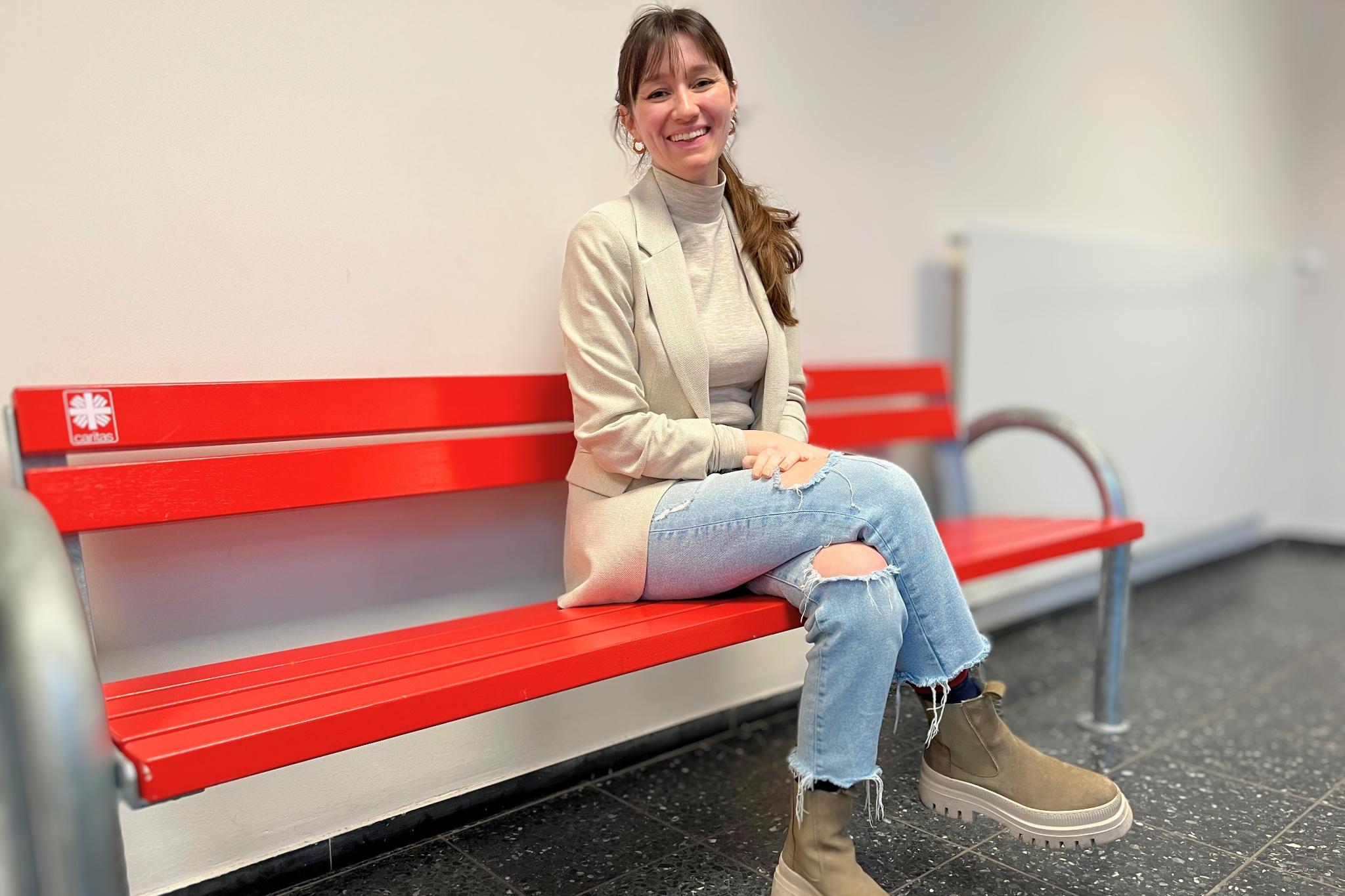 Auf dem Foto sitzt Hannah Matthews auf einer roten Bank mit einem kleinen Caritas-Logo. Sie trägt Jeans, einen hellen, beigefarbenen Pulli und eine Jacke in fast gleicher Farbe. Der Kopf ist nach rechts zur Kamera gewendet, die langen braunen Haare fallen in einem Pferdeschwanz über die linke Schulter, sie lächelt.
