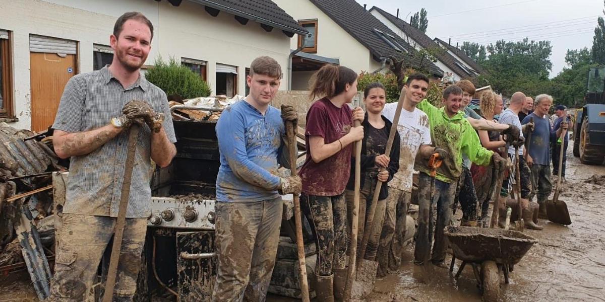 Das Foto wurde im Ahrtal aufgenommen zeigt eine verschlammte Straße, im Hintergrund Ein-Familien-Häuser, rechts am Bildrand ein Radlader. Im Mittelpunkt etwa ein Dutzend Menschen, die in einer Reihe nebeneinanderstehen. Alle haben Schaufeln in der Hand, sind schlammverdreckt, wirken erschöpft, lächeln aber; es handelt sich wohl um freiwillige Helfer.