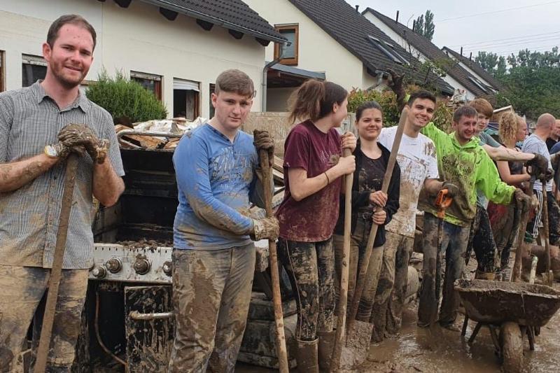 Das Foto wurde im Ahrtal aufgenommen zeigt eine verschlammte Straße, im Hintergrund Ein-Familien-Häuser, rechts am Bildrand ein Radlader. Im Mittelpunkt etwa ein Dutzend Menschen, die in einer Reihe nebeneinanderstehen. Alle haben Schaufeln in der Hand, sind schlammverdreckt, wirken erschöpft, lächeln aber; es handelt sich wohl um freiwillige Helfer.