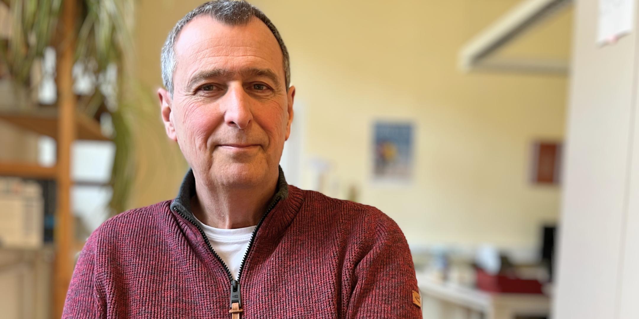 Ein älterer Mann (es handelt sich um Hubertus Kesselheim) mit kurzen grauen Haaren und freundlichem Gesichtsausdruck sitzt in einem Büro und schaut direkt in die Kamera. Er trägt einen rotbraunen Strickpullover mit Reißverschluss über einem weißen T-Shirt. Im Hintergrund sind unscharf ein Schreibtisch, Pflanzen und Büromaterialien zu sehen. (Alt-Text erstellt mit Unterstützung von KI)