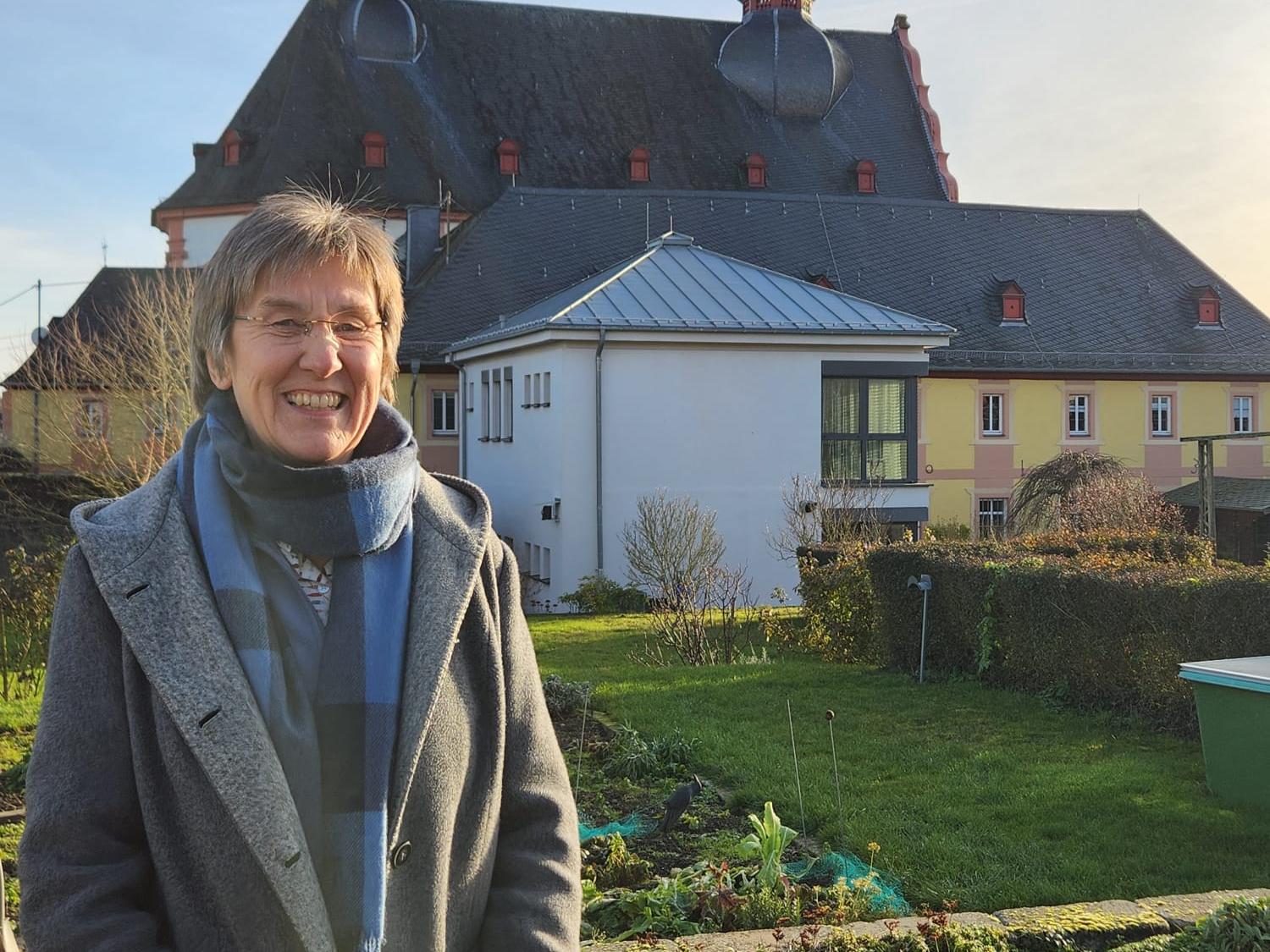 Frau steht in Garten vor Kirche mit Zwiebeltürmen im Sonnenschein. (Alt-Text erstellt mit KI-Unterstützung)