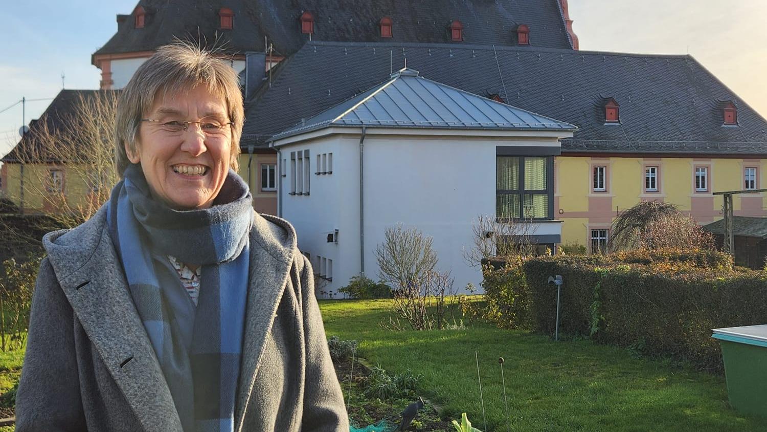 Frau steht in Garten vor Kirche mit Zwiebeltürmen im Sonnenschein. (Alt-Text erstellt mit KI-Unterstützung)