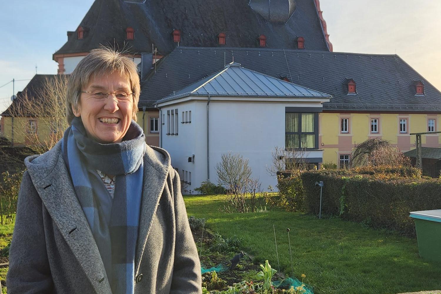 Frau steht in Garten vor Kirche mit Zwiebeltürmen im Sonnenschein. (Alt-Text erstellt mit KI-Unterstützung)