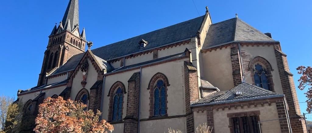 Seitlicher Blick auf eine Kirch mit spitzem Turm, die auf einem kleinen grünen Hang steht; im Vordergrund ein blühender Baum.