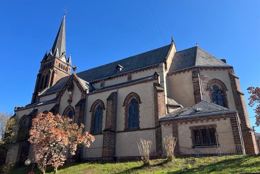 Seitlicher Blick auf eine Kirch mit spitzem Turm, die auf einem kleinen grünen Hang steht; im Vordergrund ein blühender Baum.