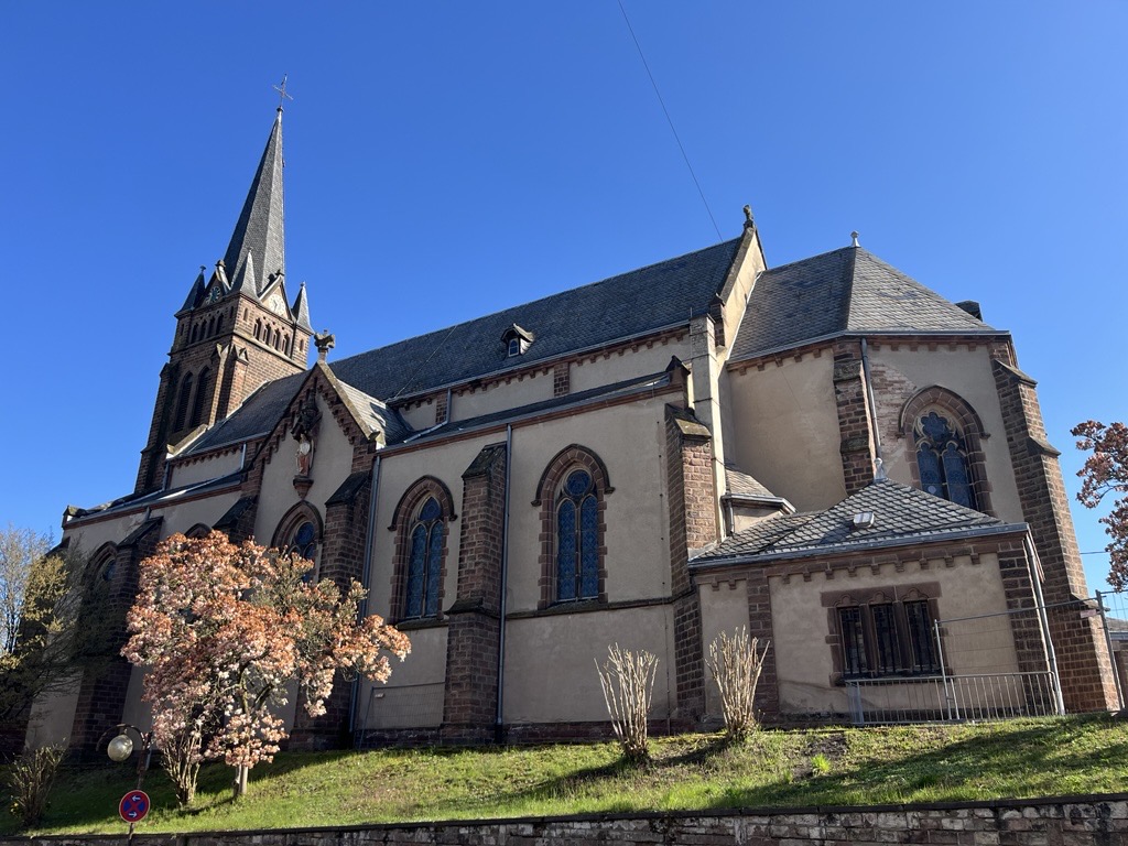 Seitlicher Blick auf eine Kirch mit spitzem Turm, die auf einem kleinen grünen Hang steht; im Vordergrund ein blühender Baum.